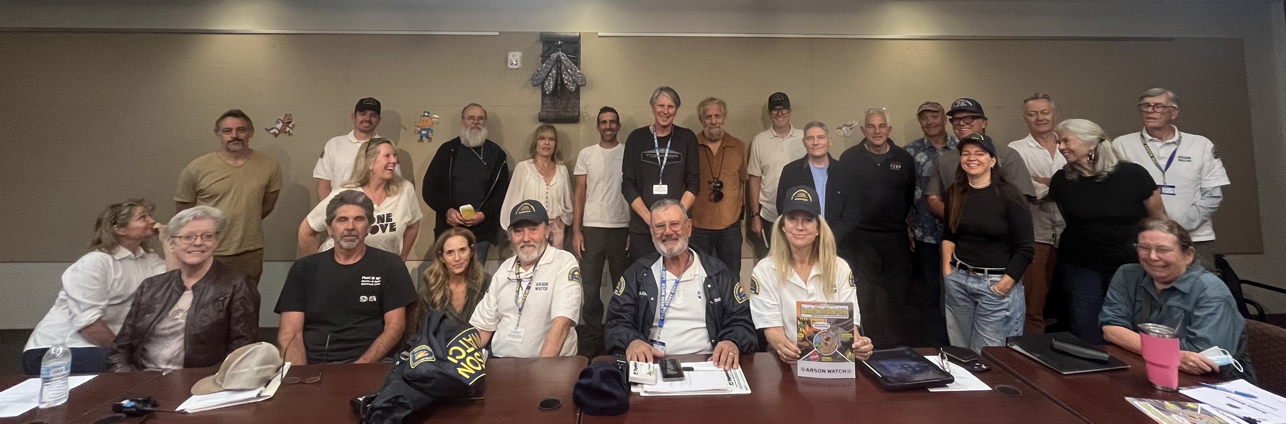 A group of around 20 people gathered together in a room for a photo, some are sitting at a table and others are standing behind them, with a variety of casual and uniformed clothing, including some with badges and caps.