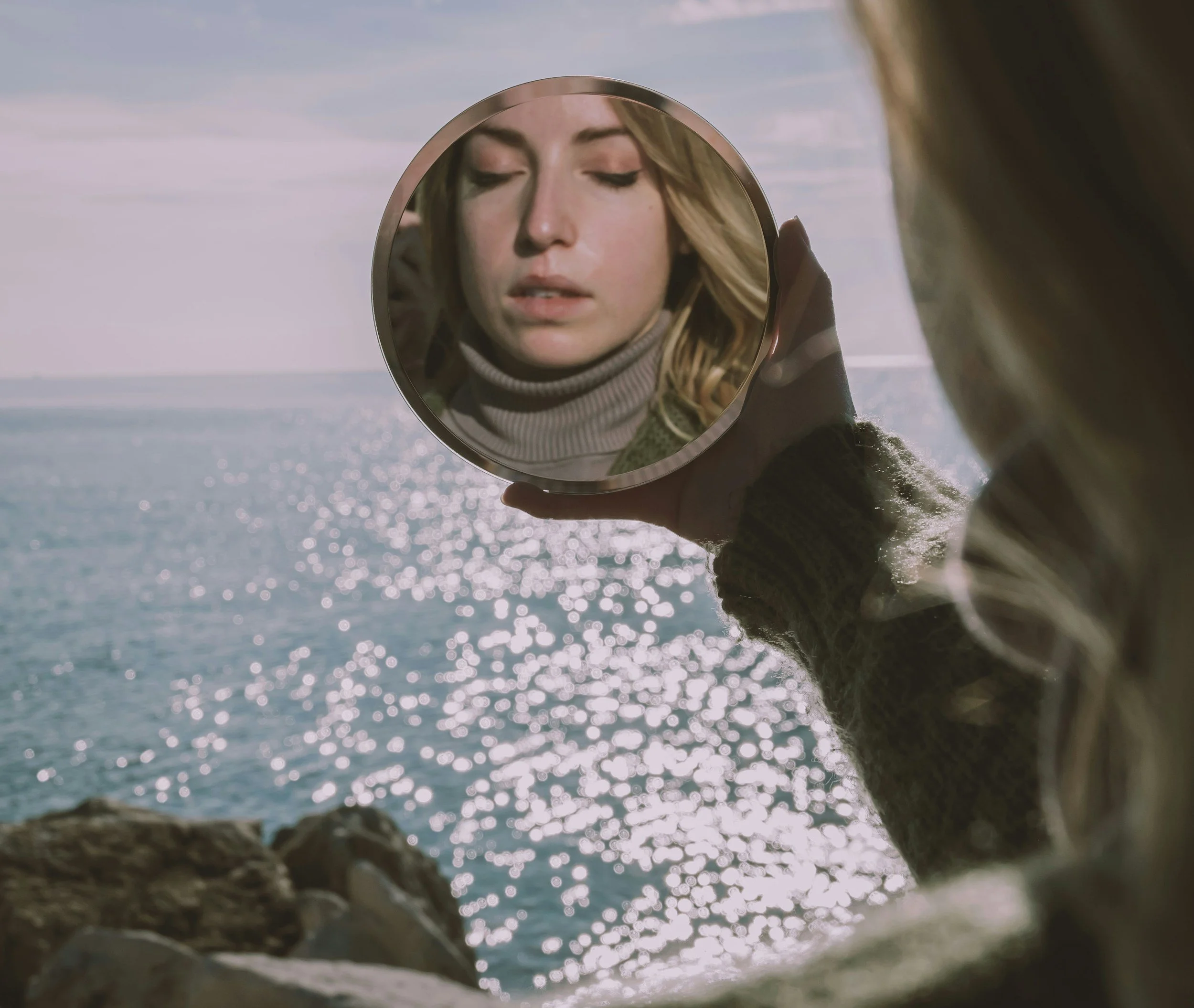 A person holding a round mirror in front of their face, reflecting a woman with blonde hair and closed eyes, by a body of water with sunlight glistening on the surface, under a sky with clouds.