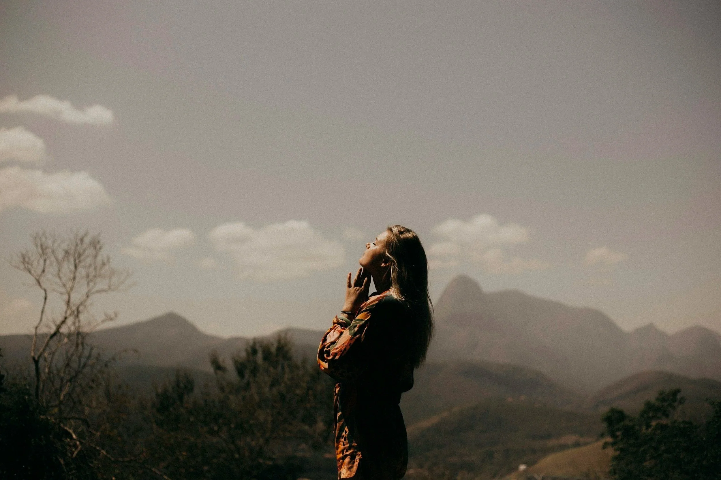 A woman standing outdoors with mountains in the background, basking in sunlight, wearing a patterned jacket and a gold wristwatch.
