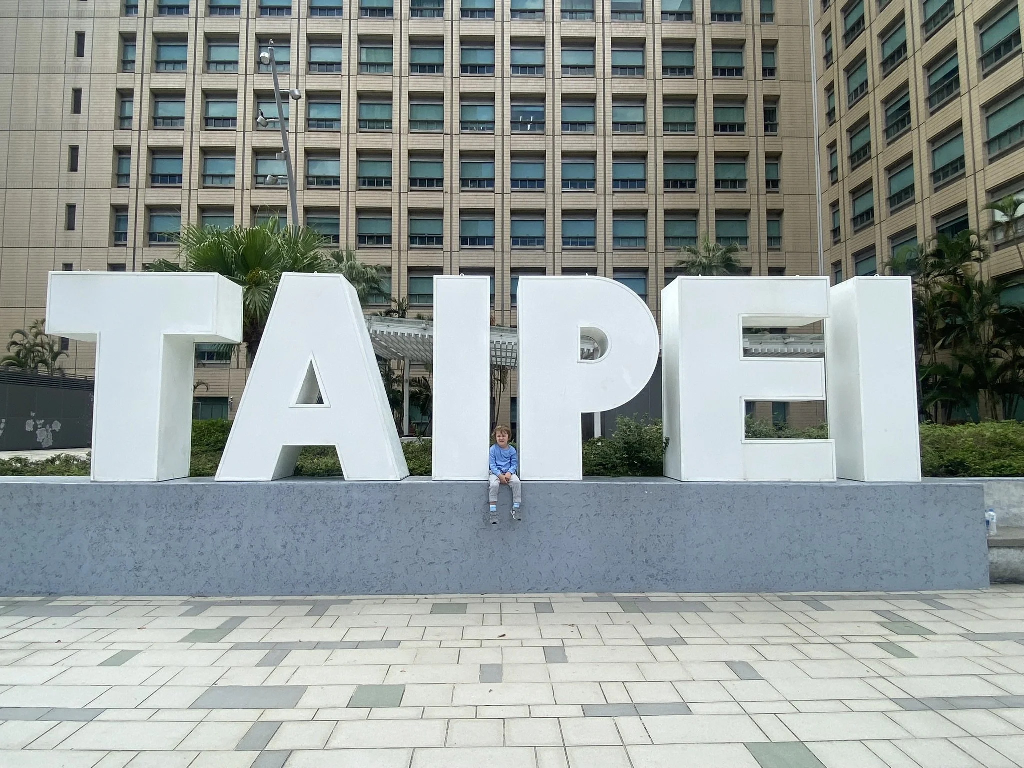A child sitting beneath large white letters spelling “TAIPEI” in a city plaza.