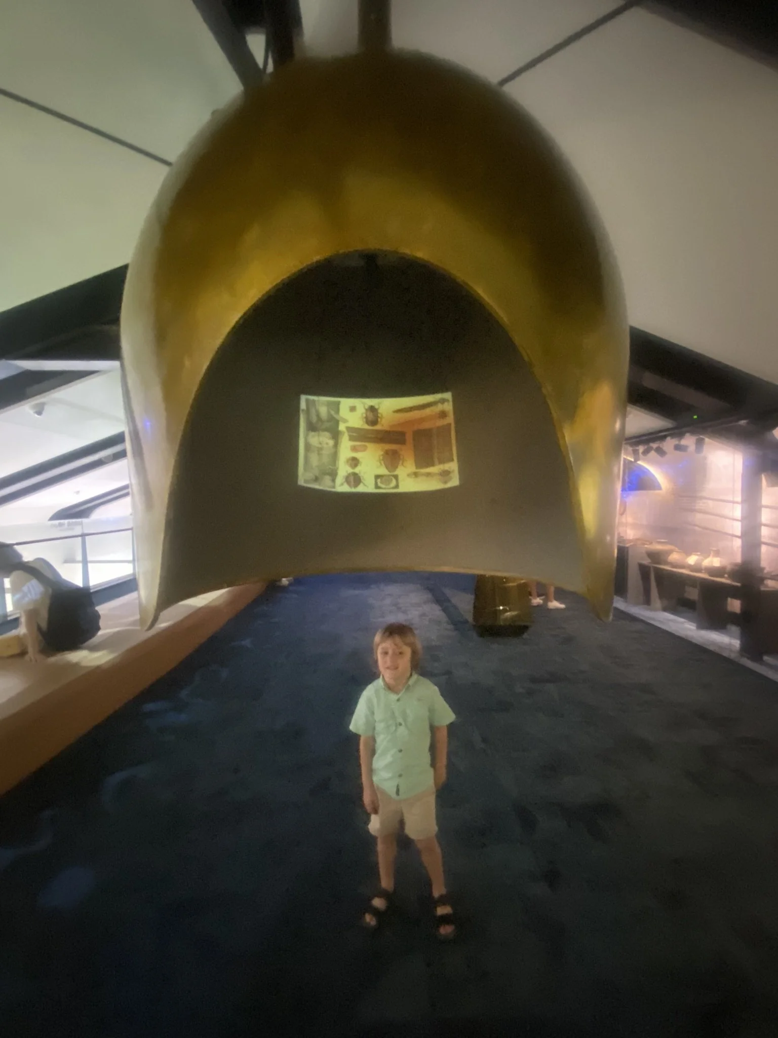 A young boy standing inside a museum under a large, curved, yellow metallic structure resembling a spacecraft or exhibit. The boy is wearing a light green shirt, beige shorts, and black sandals. The background includes display cases and museum lighting.