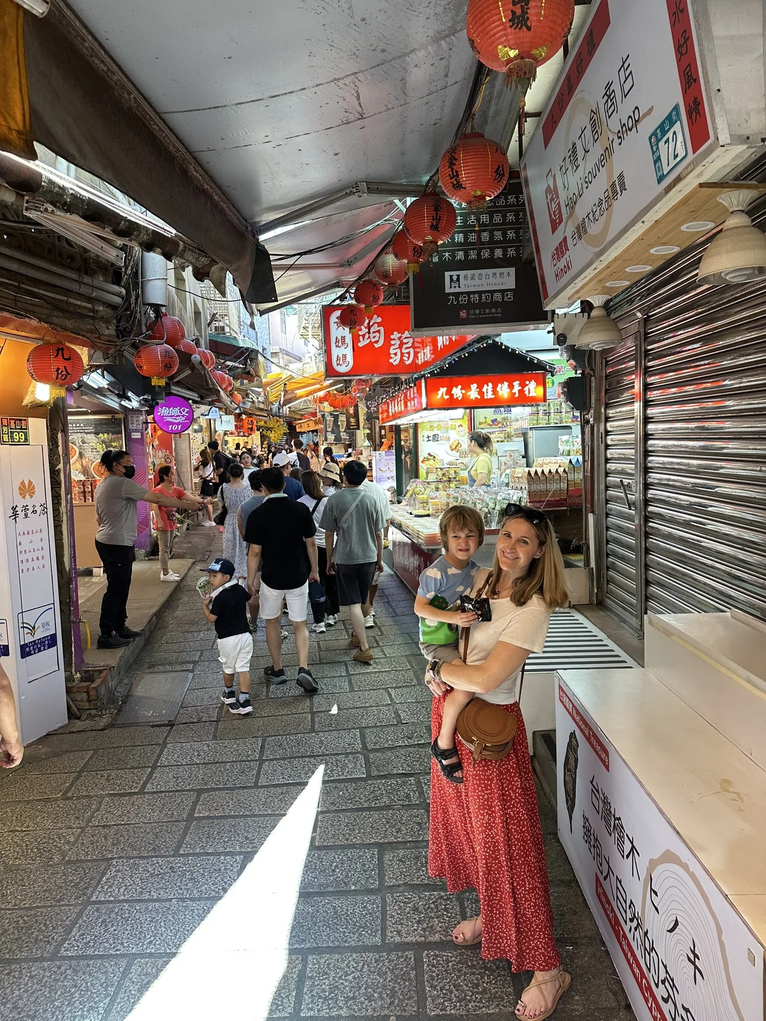 An adult holding a child on Jiufen Old Street, with shops and red lanterns behind them.