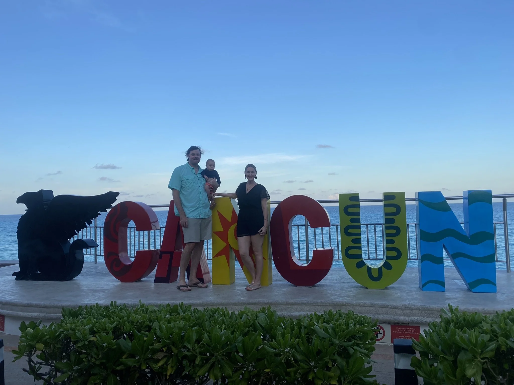 Family of three posing behind a colorful Cancún sign with the sea in the background.