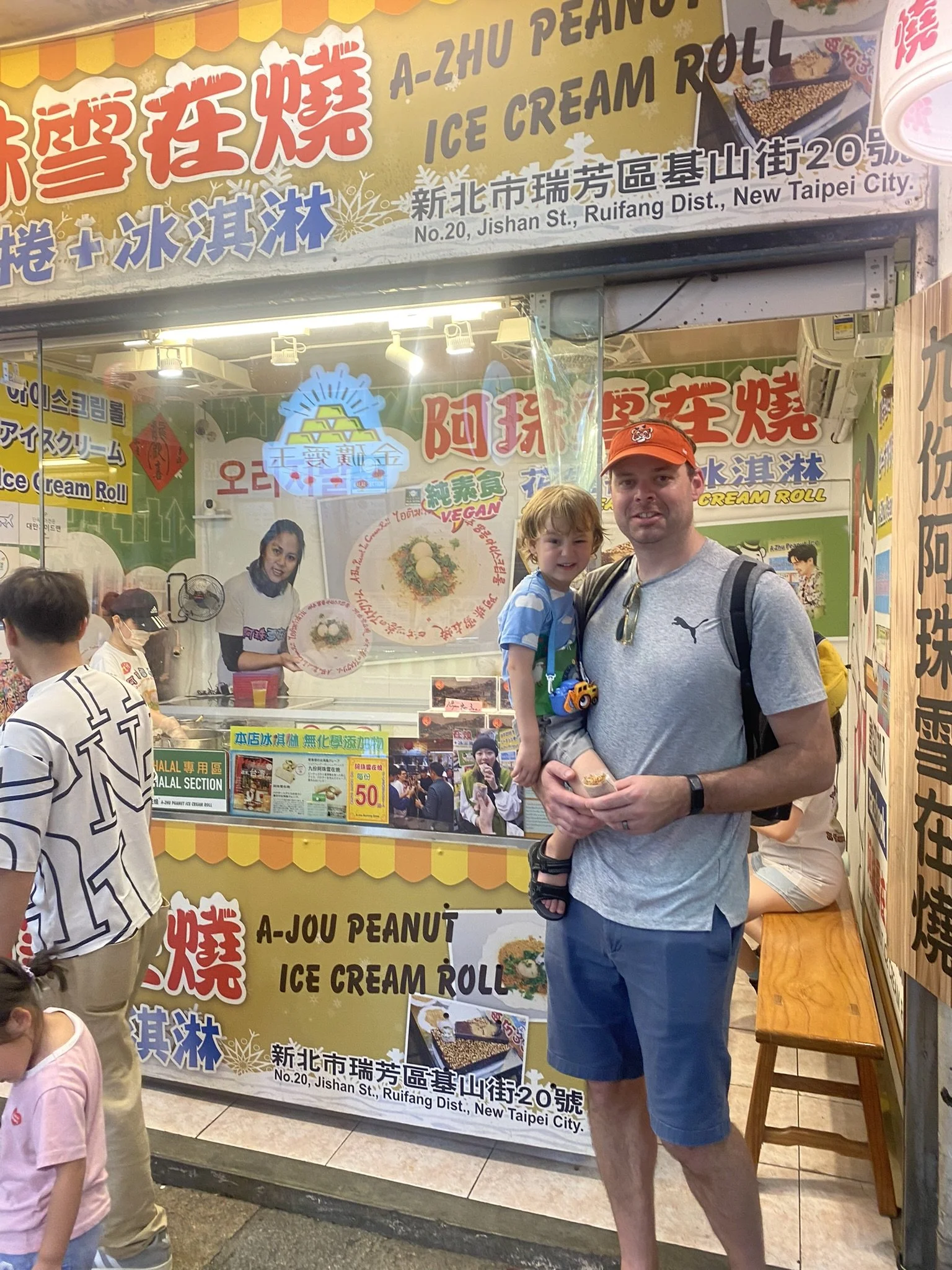 An adult holding a child in front of a peanut ice cream roll stand in Jiufen.