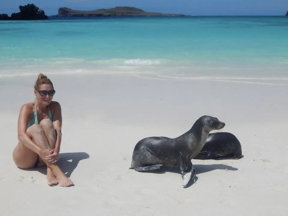 A woman sitting beside two sea lions on a white-sand beach in the Galápagos Islands.