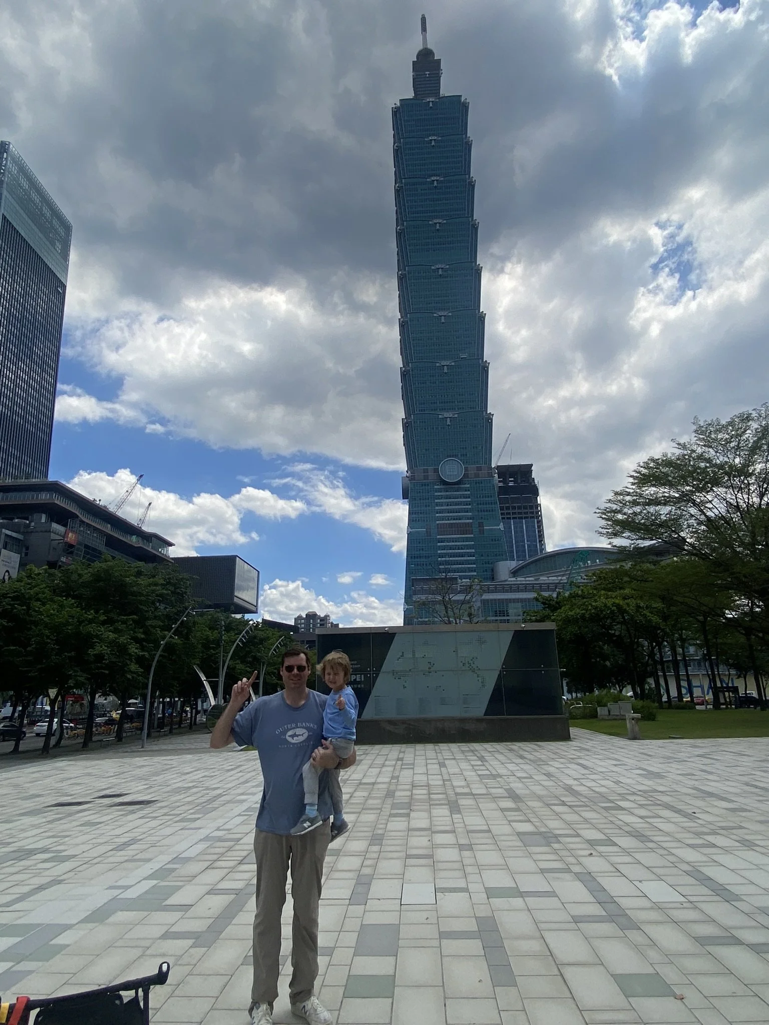 A man holding a child walking in an urban plaza with Taipei 101 skyscraper in the background, partly cloudy sky, and trees on sides.