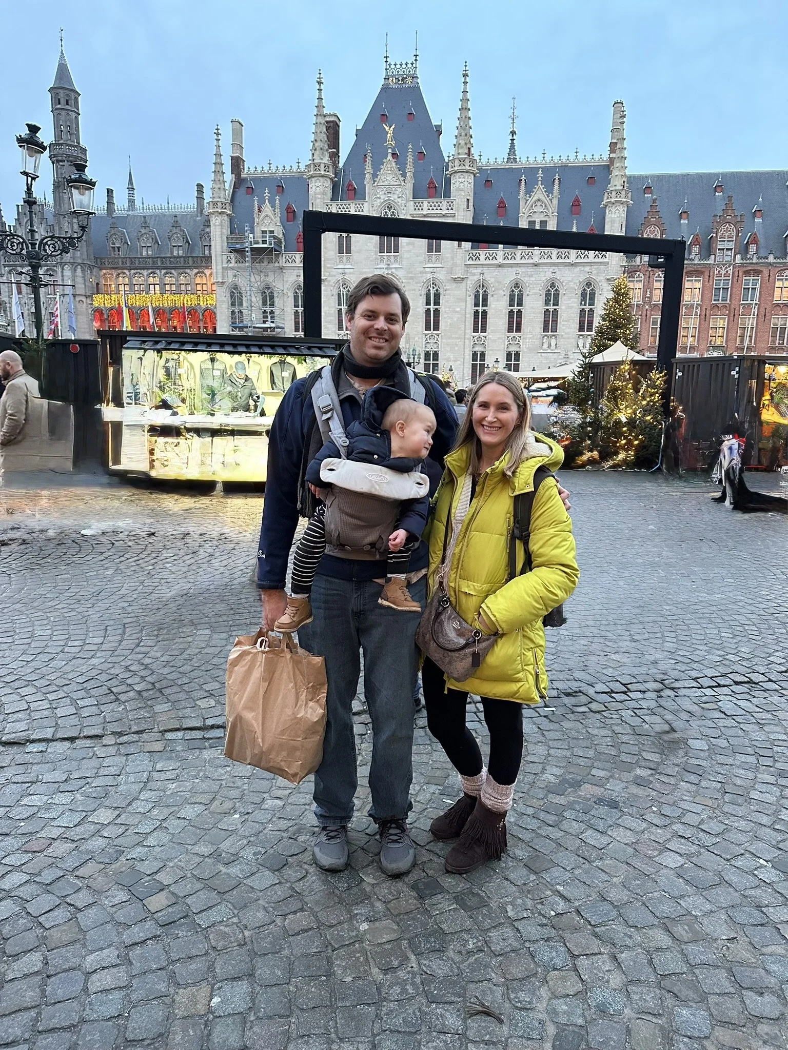A family of three standing in a cobblestone square in Bruges, with historic buildings behind them.