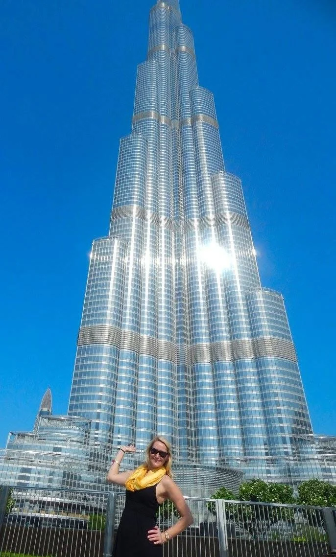 A woman posing in front of the towering Burj Khalifa skyscraper in Dubai, with a clear blue sky overhead.