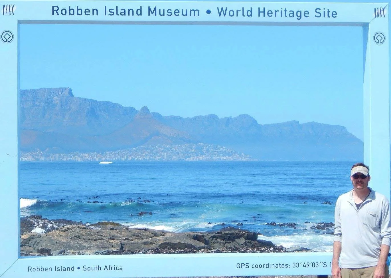 A man standing on rocky shoreline at Robben Island Museum, South Africa, with Table Mountain in the background, and a blue informational frame around the scene.