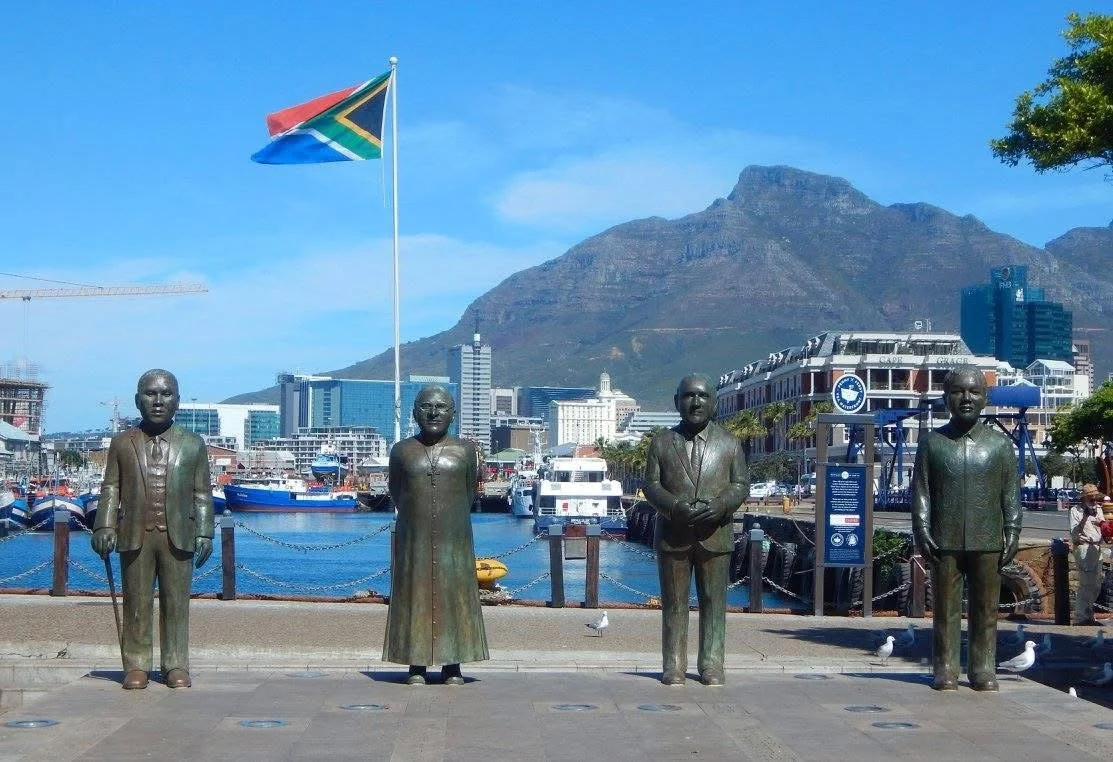 Four bronze statues beneath a South African flag, with Table Mountain behind them.