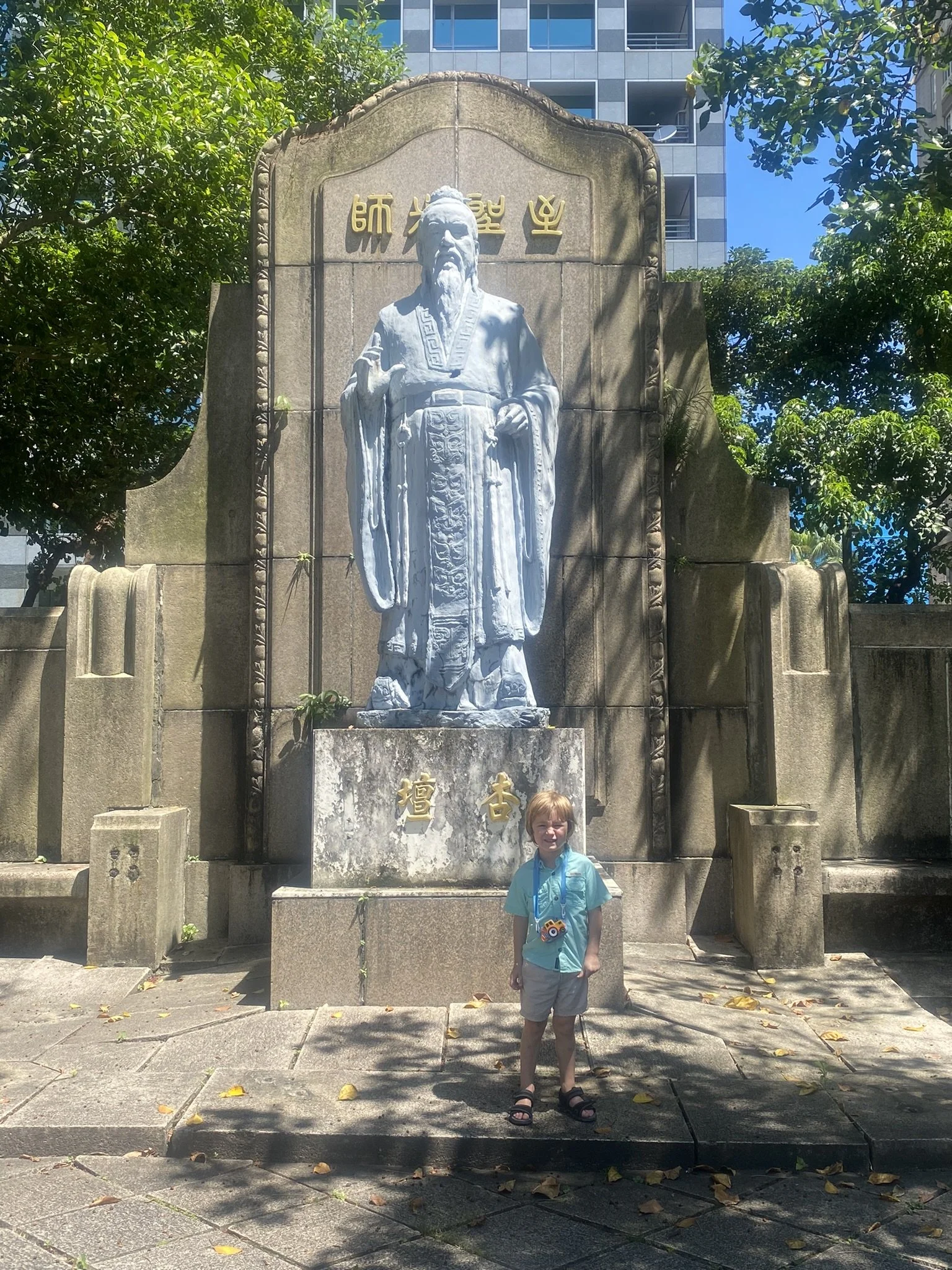 A child with a camera standing in front of a large stone statue in Taipei.
