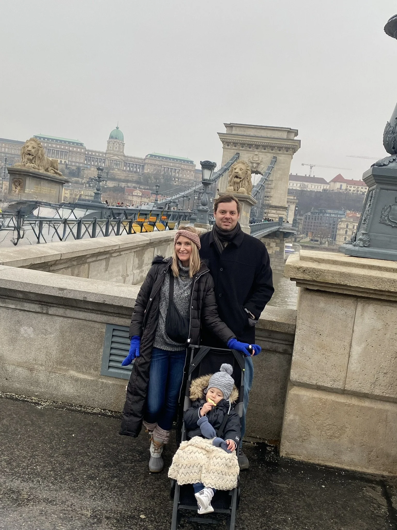 A family of three beside the Danube in Budapest, with the Chain Bridge and historic buildings behind them.