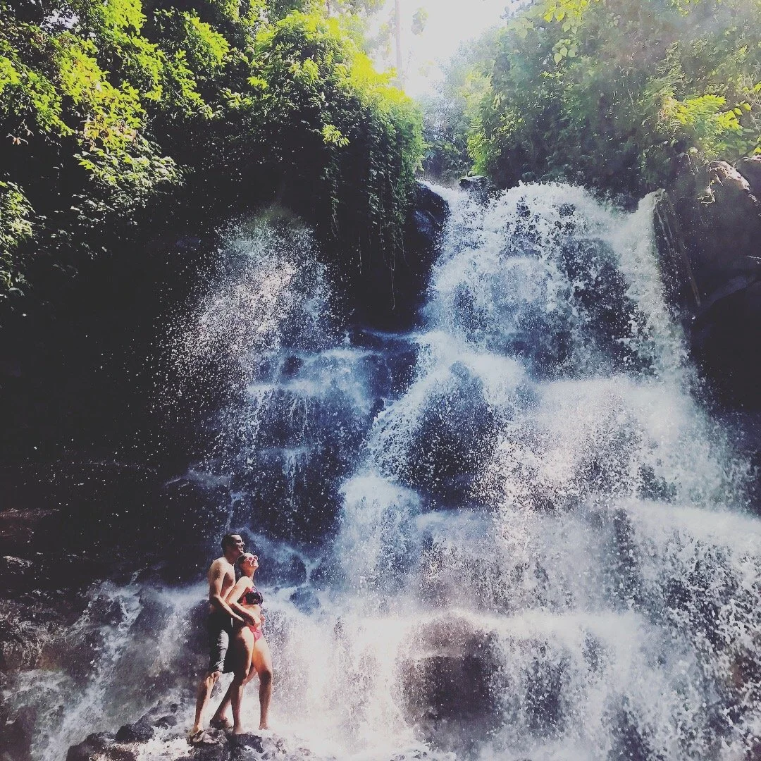 Couple standing on rocks at the base of a waterfall near Ubud, Bali.