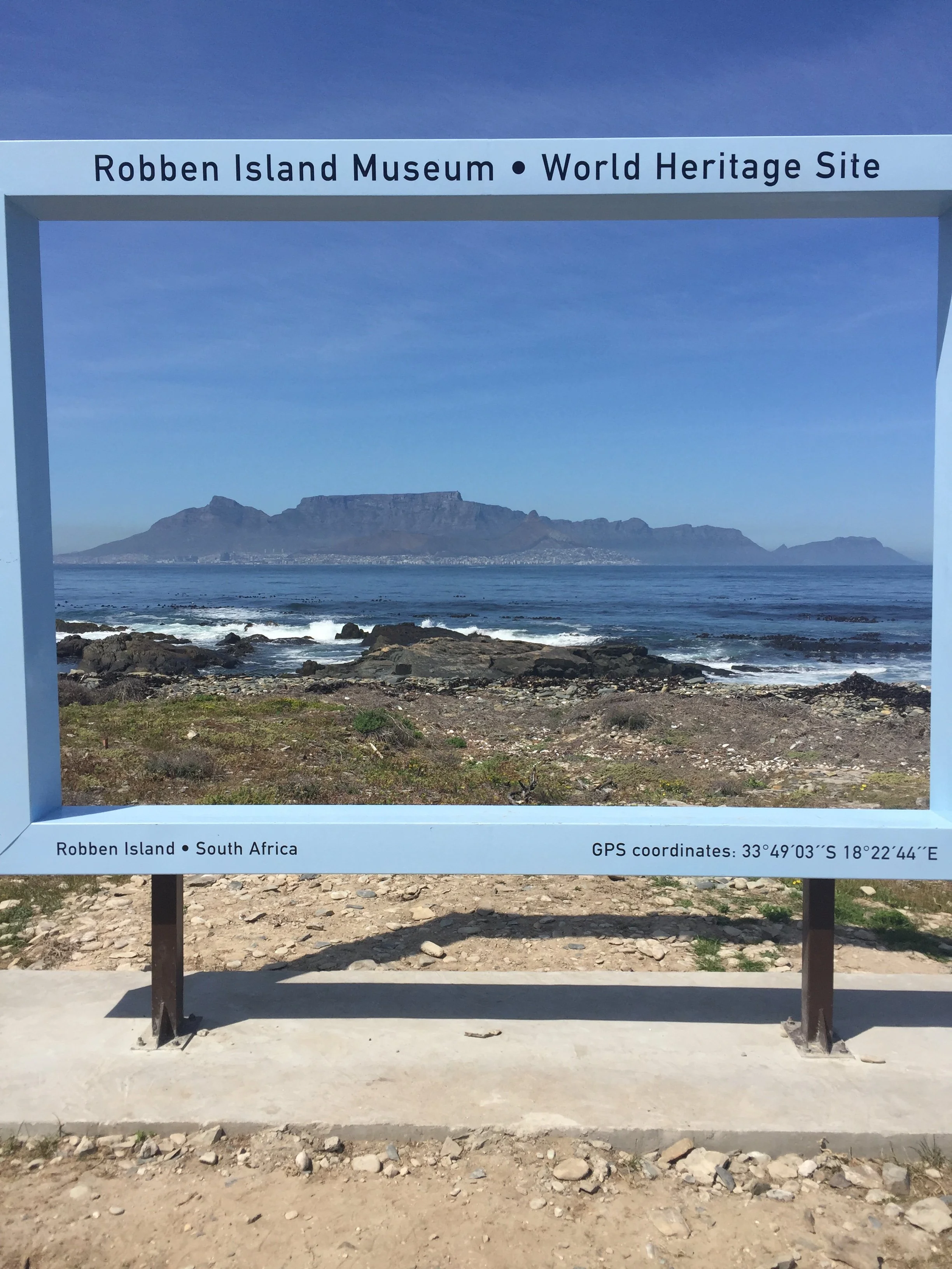 Signboard with a view of Robben Island off the coast of South Africa, with mountains, ocean, and rocks in the background.