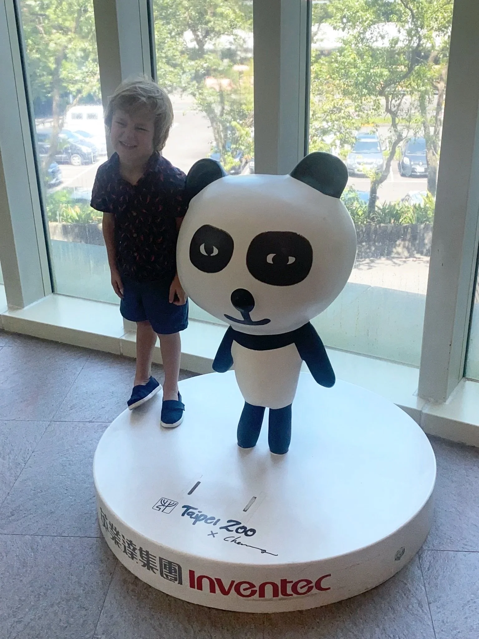A young boy standing next to a panda mascot statue inside a building near a large window with a parking lot and trees outside. The panda is black and white with a rounded head and large black eyes, and the boy is smiling and wearing a dark shirt with a pattern and blue shorts.