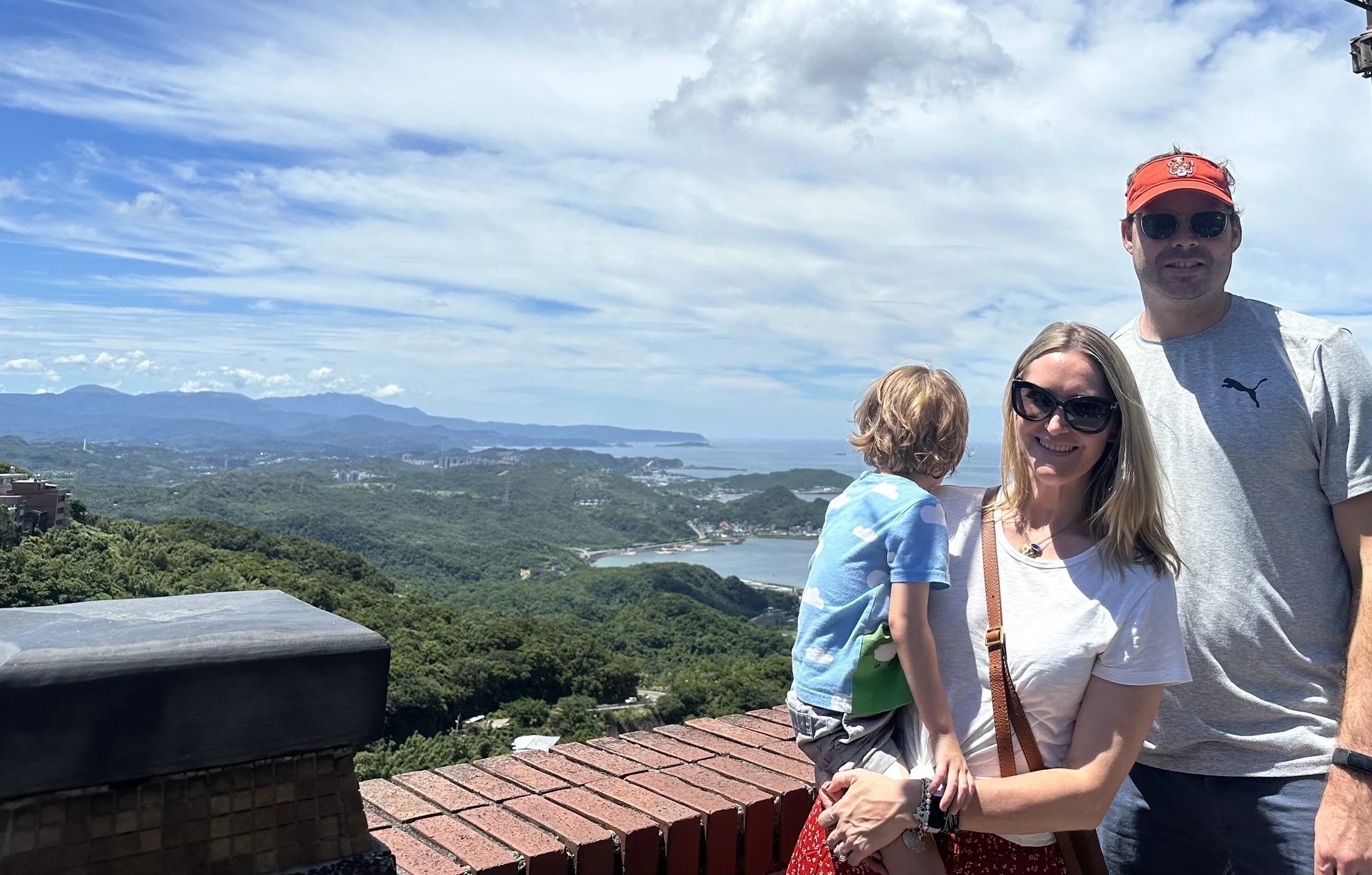 Two adults and a child at a Jiufen overlook, with mountains and water in the distance.