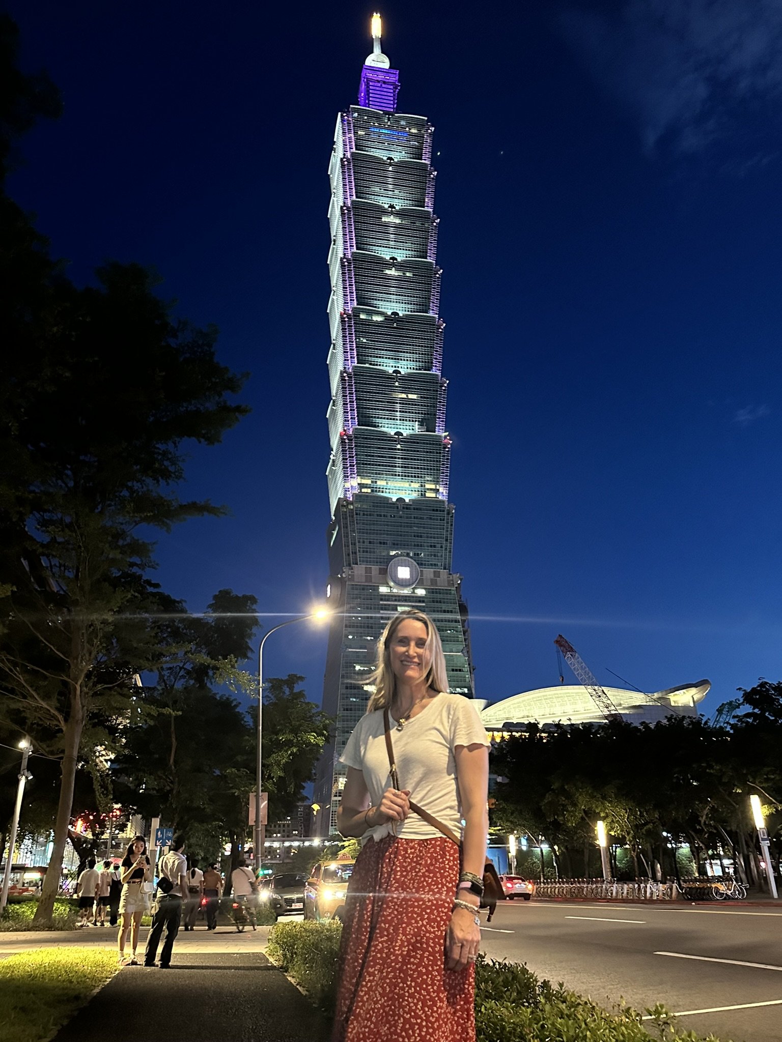 A woman with blonde hair wearing a white top and a red patterned skirt stands on a city street at night, with the Taipei 101 skyscraper illuminated behind her. The sky is dark and the street is busy with people and cars.