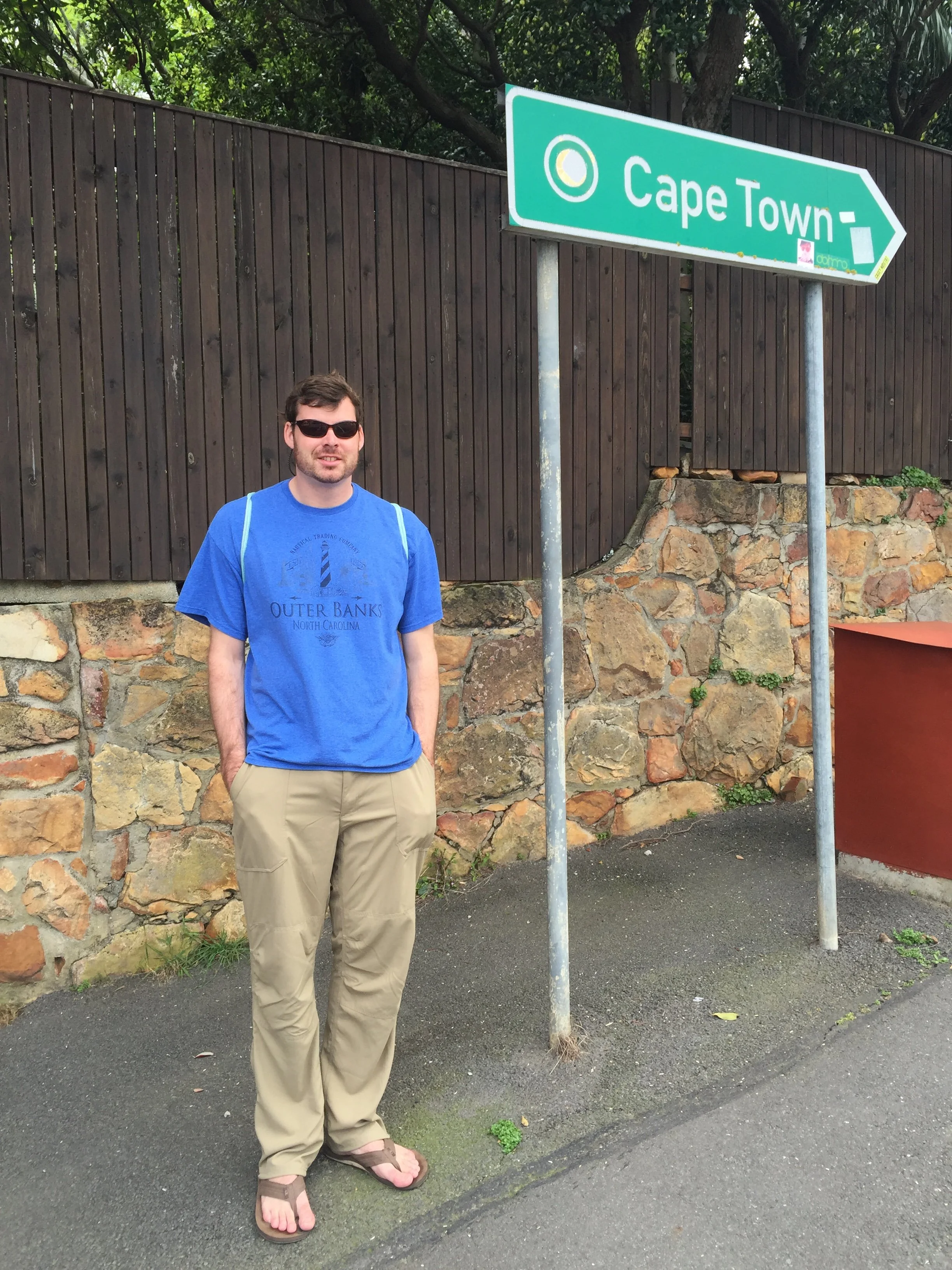 A man in sunglasses, a blue T-shirt, beige pants, and flip-flops standing on a sidewalk next to a street sign that points to Cape Town. There is a stone wall and a wooden fence behind him, and trees overhead.