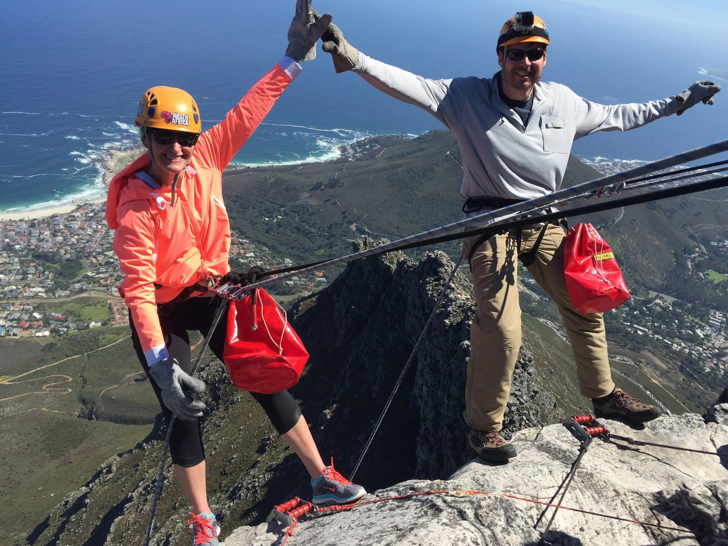 Two people in helmets high-fiving atop Table Mountain before abseiling, with Cape Town’s coast behind them.