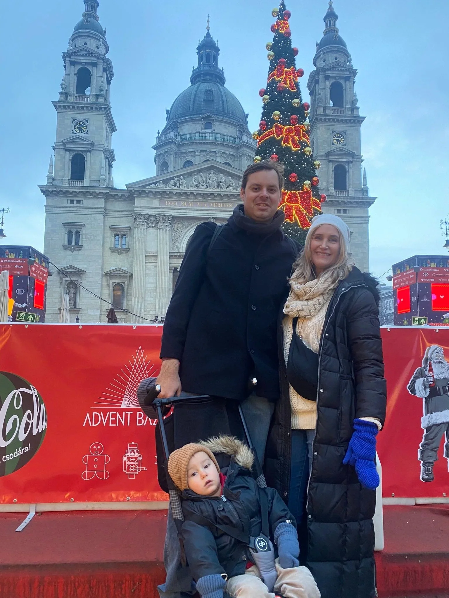 Family with a child in a stroller beside a Christmas tree in front of St. Stephen’s Basilica in Budapest.