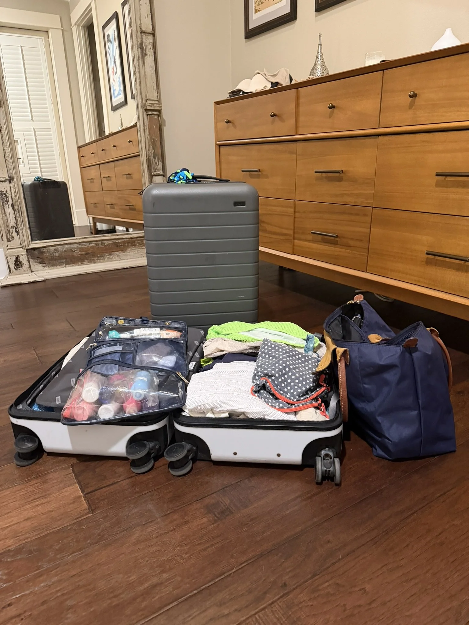 Open suitcase filled with toiletries and clothes on wooden floor, surrounded by bags, with a wooden dresser and framed artwork in the background.