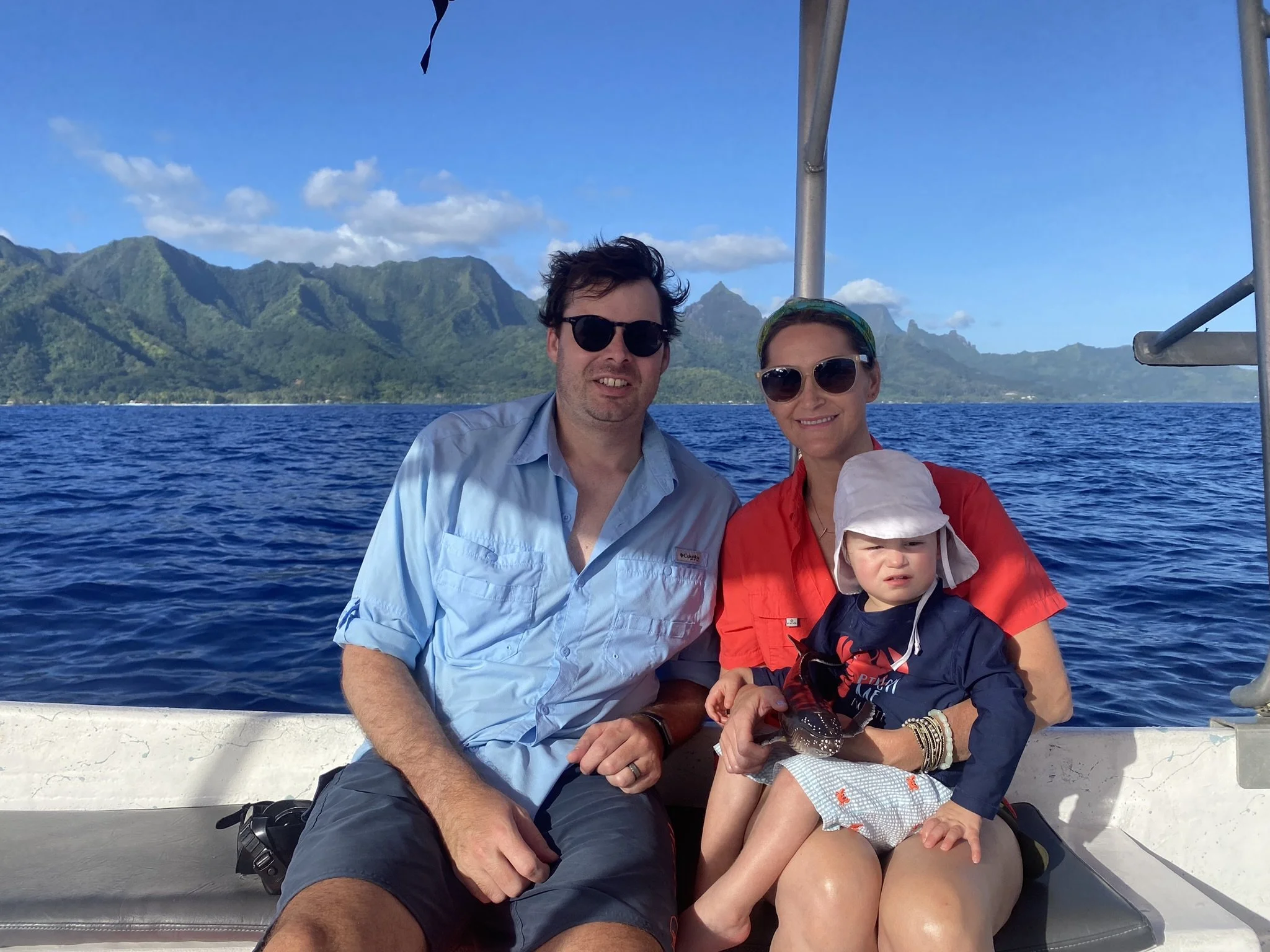 A family of three on a boat off Moorea, with green mountains behind them.