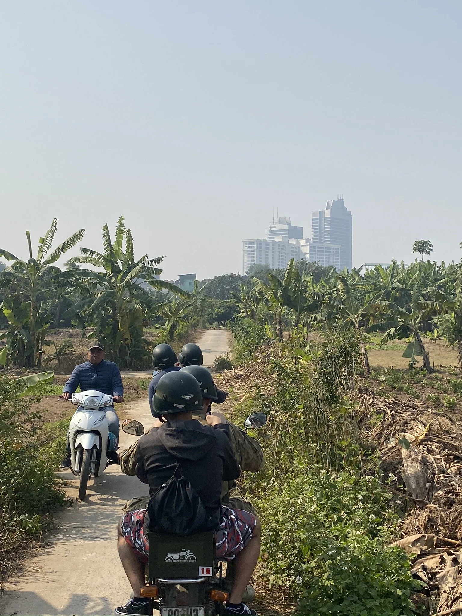 Motorcyclists riding along a narrow path through banana fields in Hanoi.