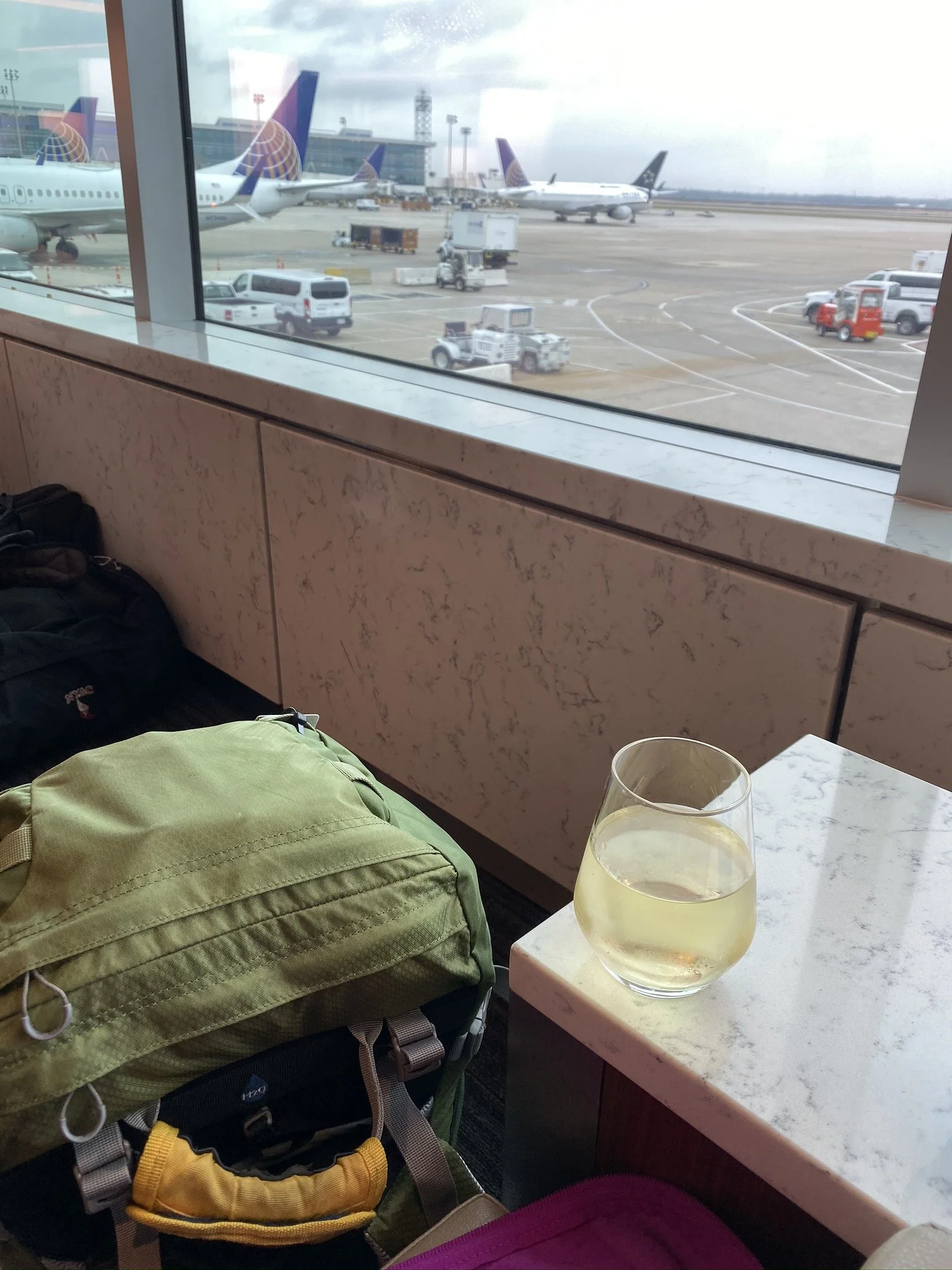 View of aircraft and ground vehicles through an airport lounge window, with a drink and travel bags on the counter.