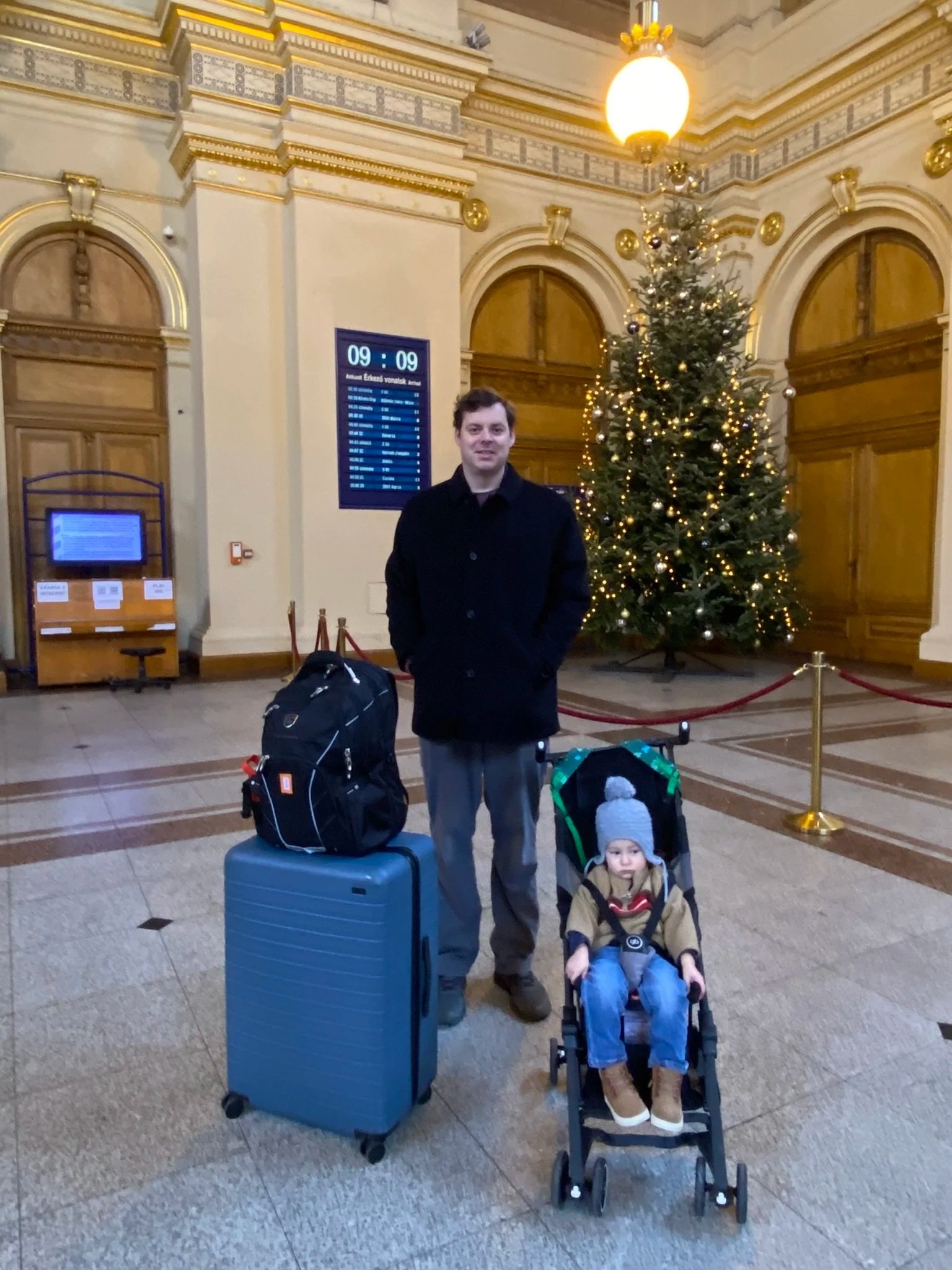 Father and young son with a stroller and suitcase beside a Christmas tree in a Budapest train station.