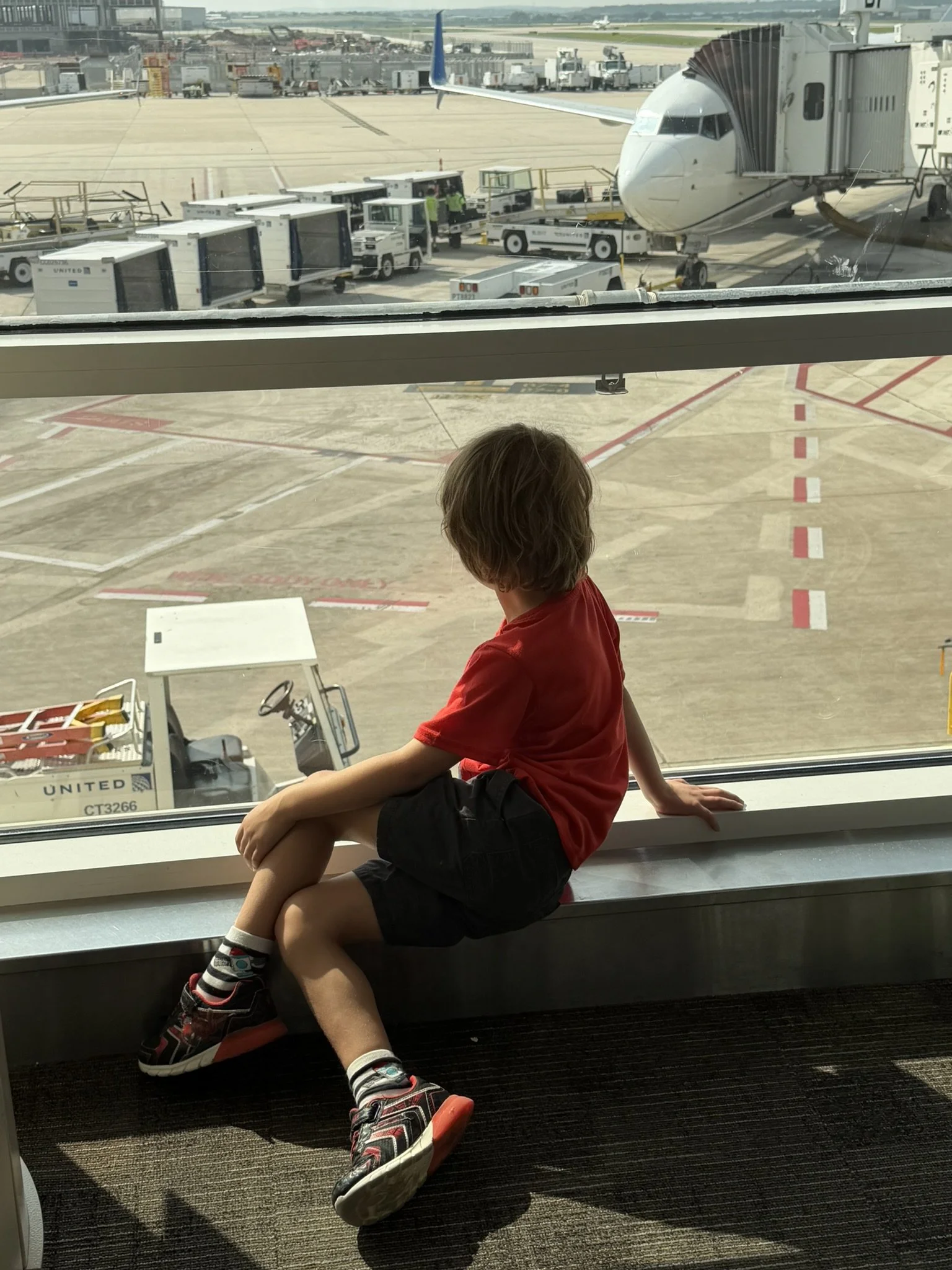 A young boy looking out the window at a parked airplane with a jet bridge attached, airport ground crew and vehicles nearby.