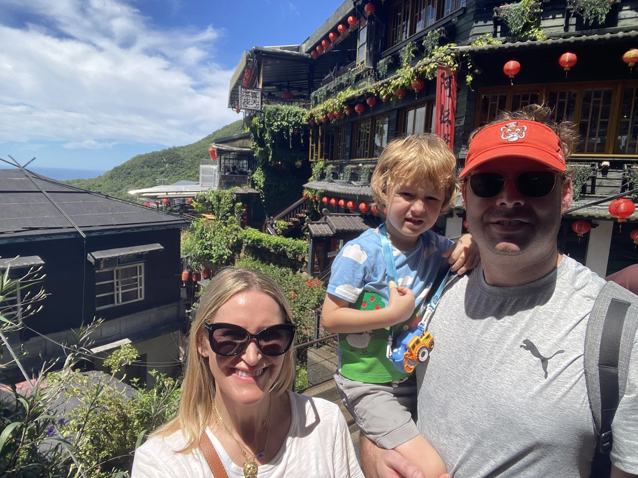 Two adults and a child in Jiufen, with hillside buildings and red lanterns behind them.