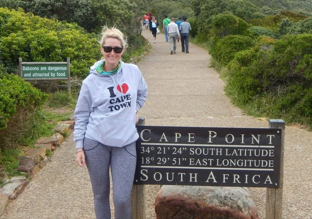 A woman wearing a gray hoodie with 'I love Cape Town' written on it, standing next to a sign indicating Cape Point in South Africa, with other people walking along the trail.