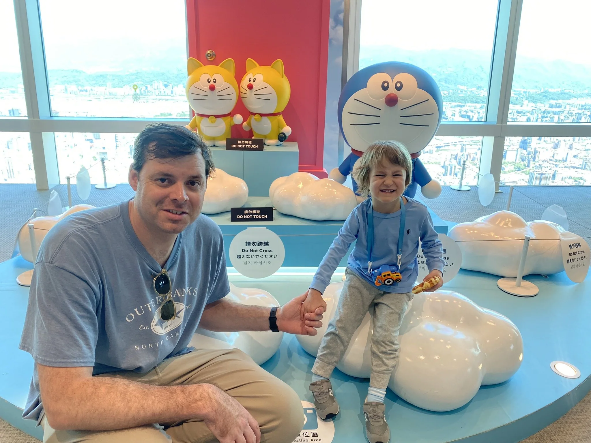 An adult and a child posing beside Doraemon figures inside Taipei 101, with the city visible through the windows.