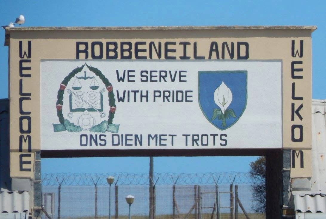 Welcome sign for Robben Island with the slogan "We serve with pride" and the Afrikaans translation "Ons dien met trots." The sign includes a logo with a book, scales, and a laurel, and another emblem with a white flower on a blue background, set against a barbed wire fence and clear blue sky.