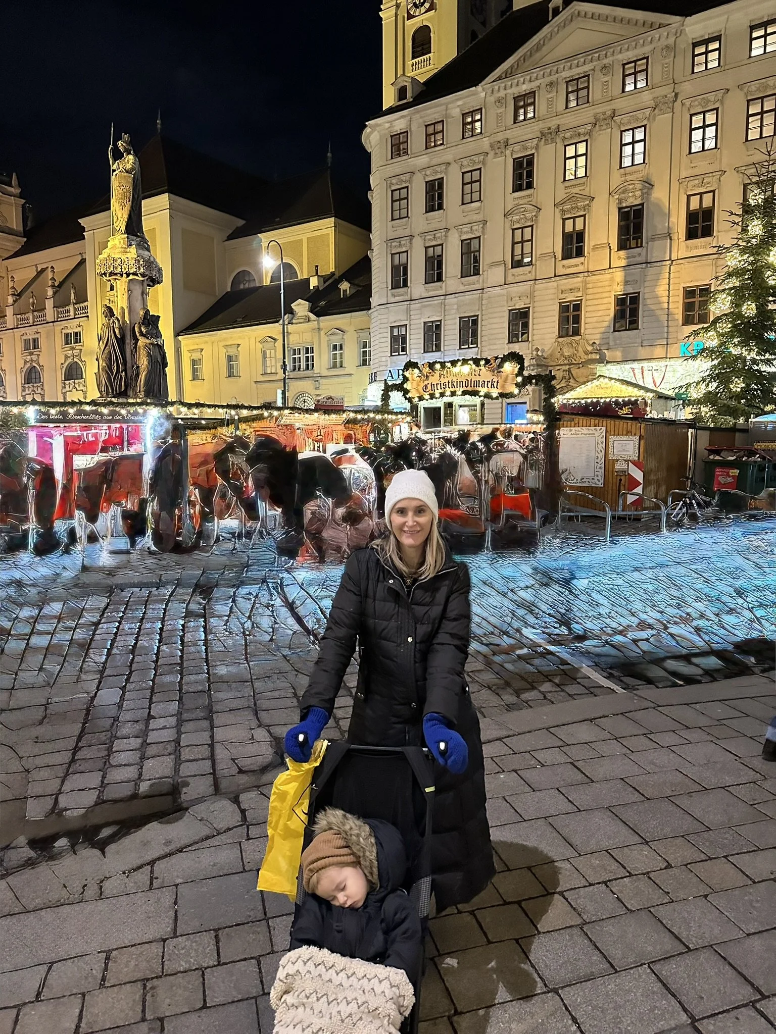 A woman stands in front of Christmas market stalls at night, with a church and historic buildings illuminated in the background. A child sleeps in a stroller at her feet.