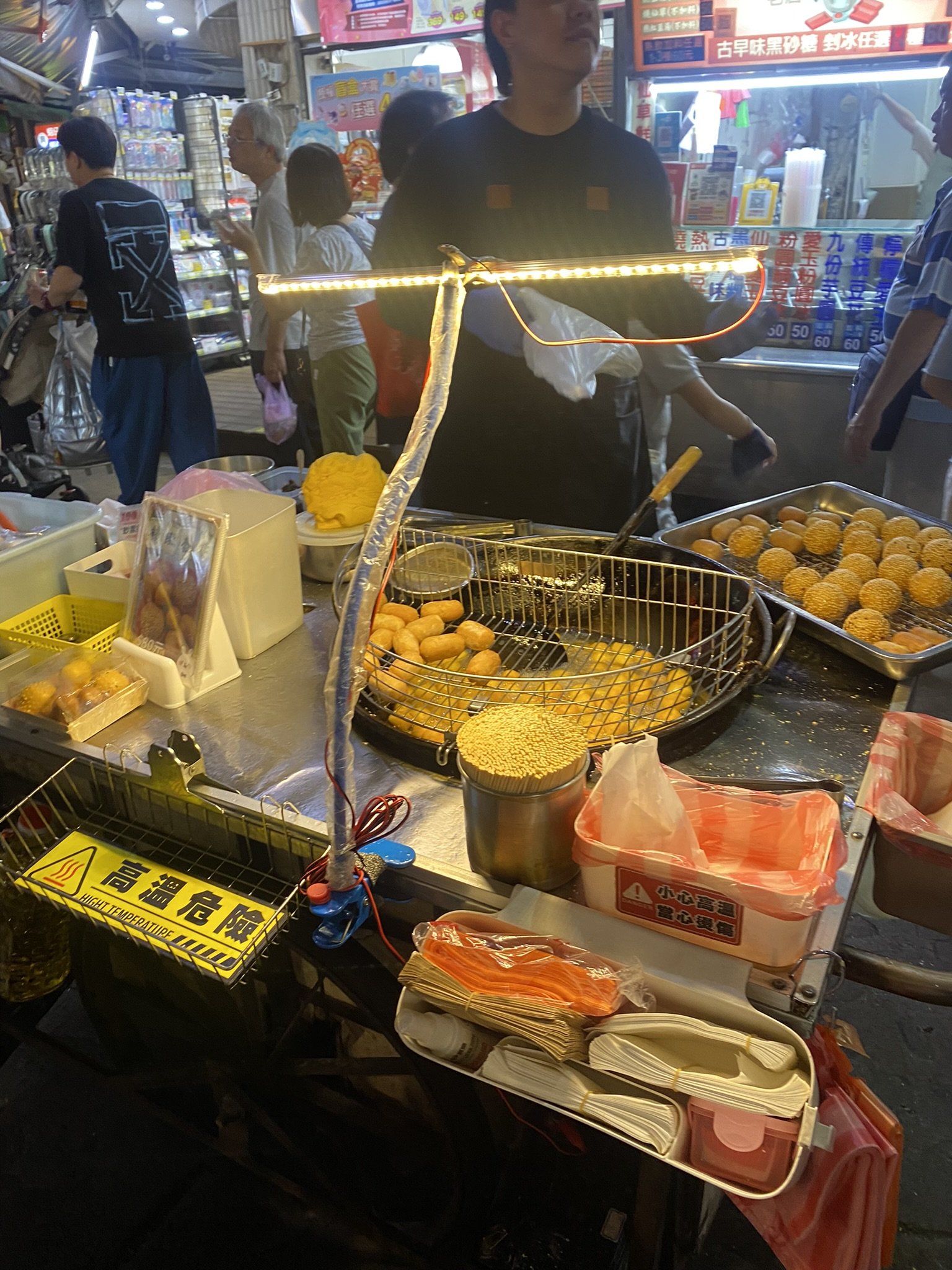 A street food stall in an Asian market at night, with various fried snack items in baskets, utensils, and napkins, and a group of people shopping in the background.
