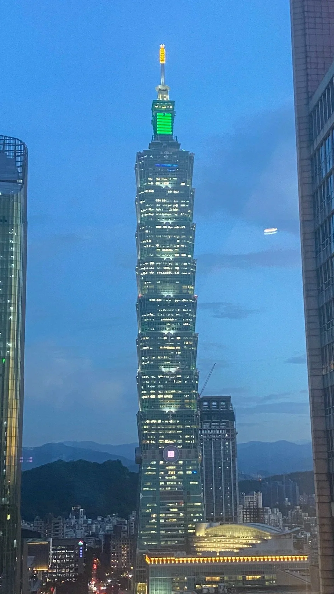 Night view of Taipei 101 skyscraper illuminated with green and yellow lights, surrounded by other tall buildings in Taipei, Taiwan, with mountains in the background.