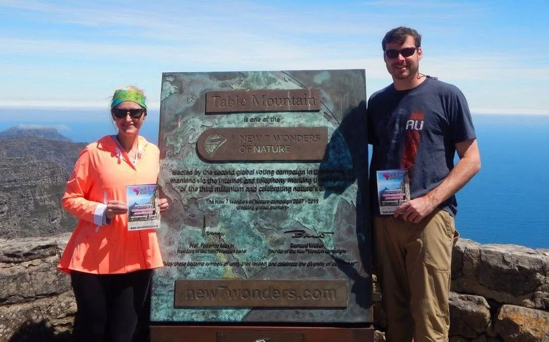 Two people standing outdoors on a mountain summit holding signs, with a large engraved sign behind them that reads 'Table Mountain is one of the New 7 Wonders of Nature,' and a view of mountains and water in the background.
