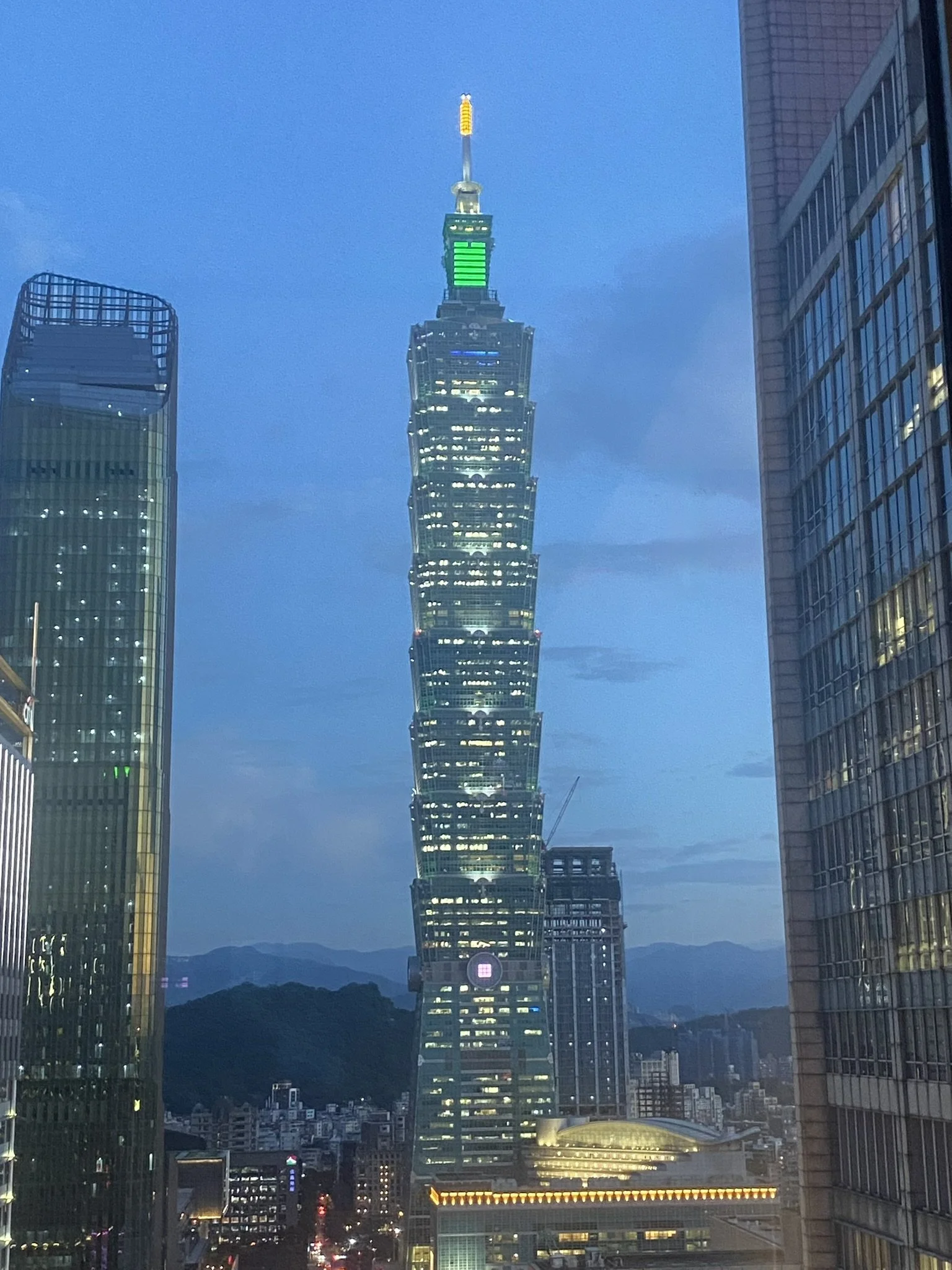Nighttime cityscape featuring the Taipei 101 skyscraper illuminated with green and yellow lights, flanked by modern glass buildings on both sides, with mountains and a blue sky in the background.