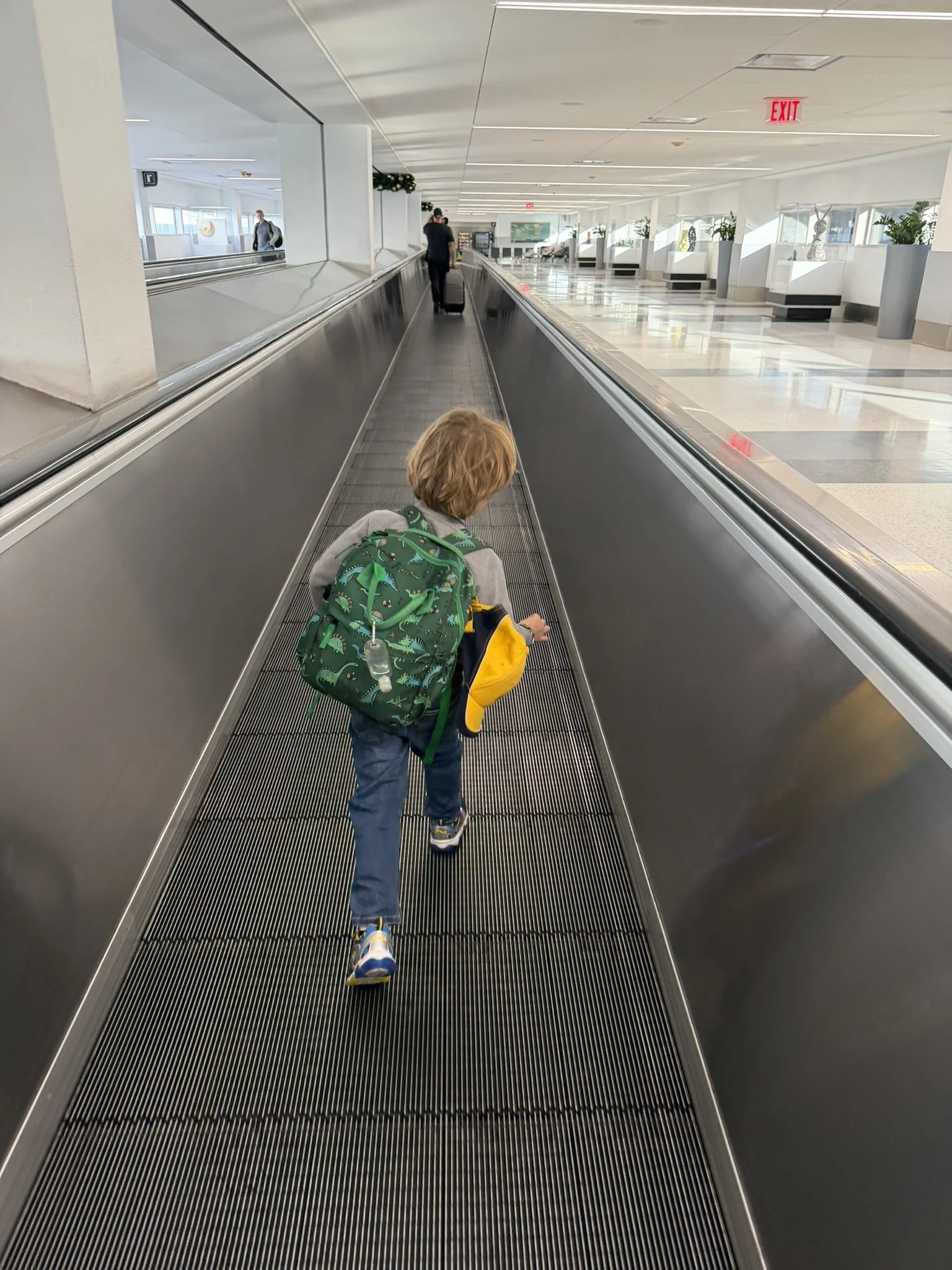 A young child with a backpack and a water bottle walking on a moving walkway at an airport, with an adult pulling a suitcase ahead in the distance.