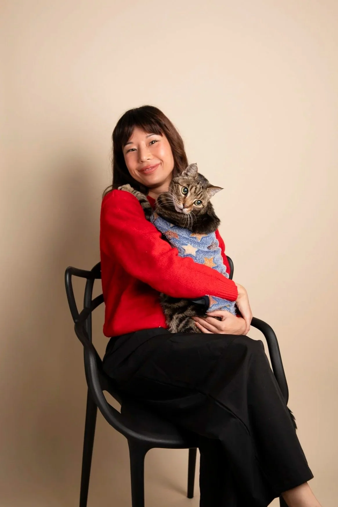 Therapist with short dark hair and a red sweater is sitting on a black chair and holding a tabby cat wearing a blue sweater. She is smiling and looking at the camera against a plain beige background.