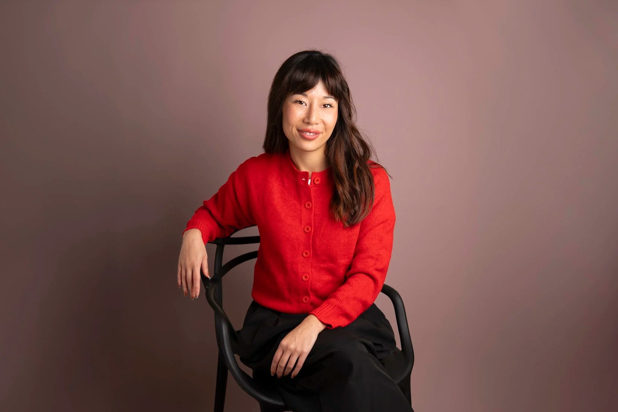 A therapist with long wavy brown hair wearing a red button-up sweater sitting on a black chair against a plain pink wall.