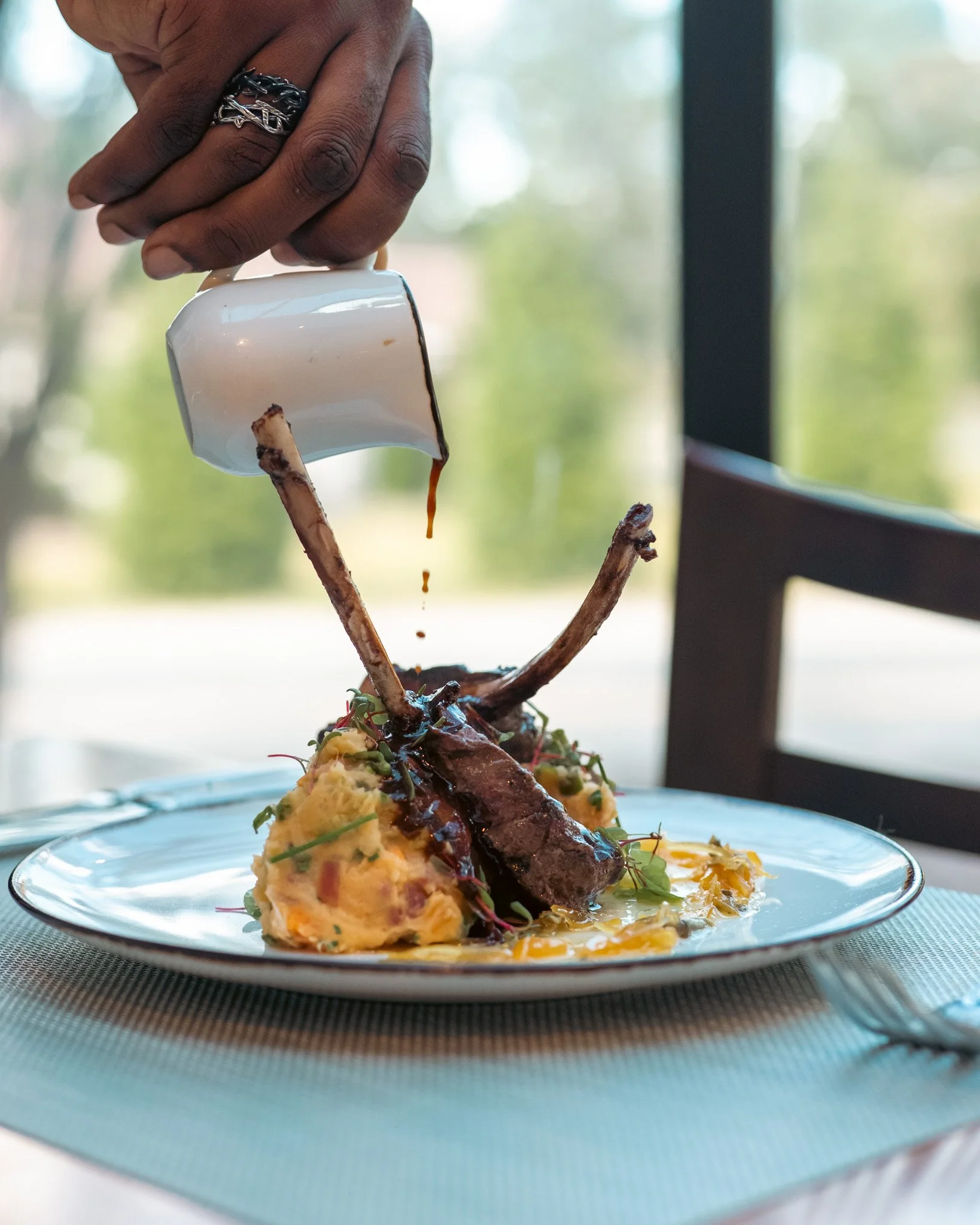 A hand with a silver ring pouring sauce over a gourmet dish featuring mashed potatoes, grilled meat, and vegetables on a white plate.