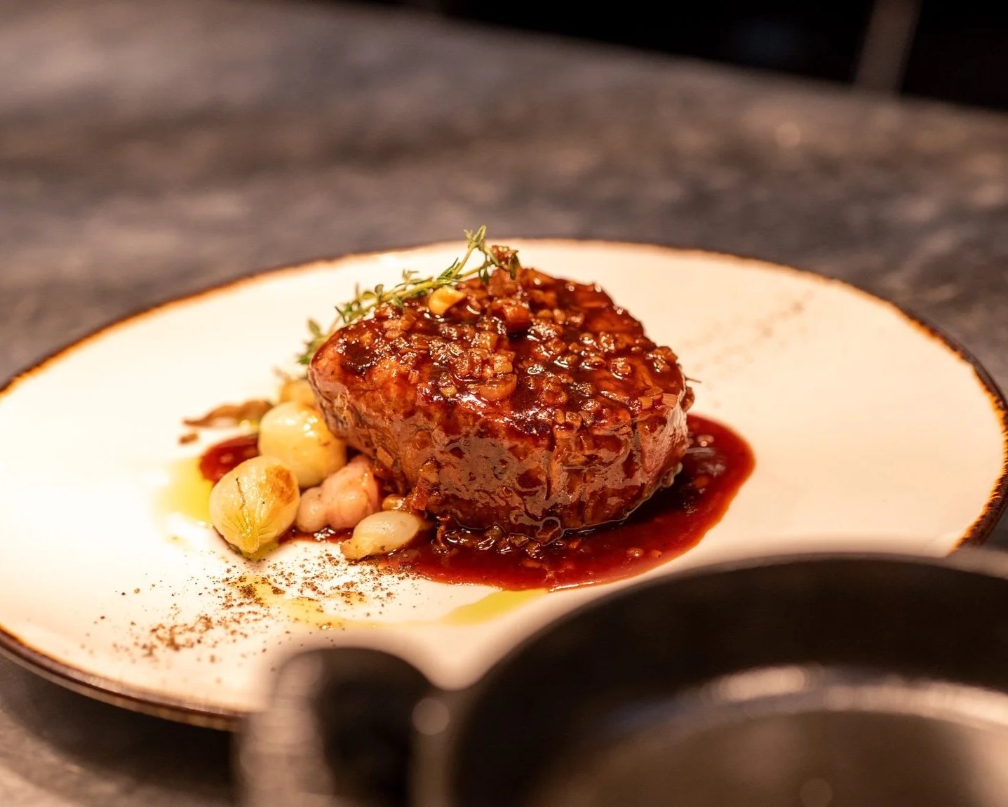 A plated gourmet entrée featuring a meatloaf glazed with sauce, garnished with herbs, accompanied by pearl onions and grape tomatoes, and finished with a sprinkle of spices.