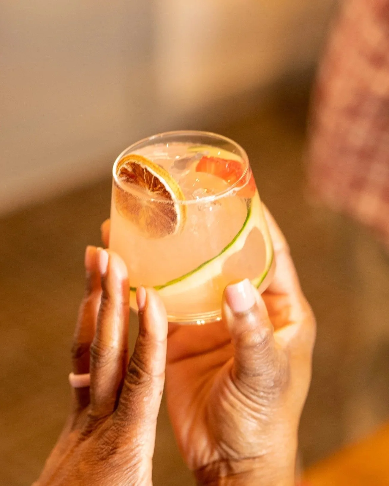 A person holding a glass of pink citrus drink with lemon and orange slices inside, and a lemon slice garnishing the rim.