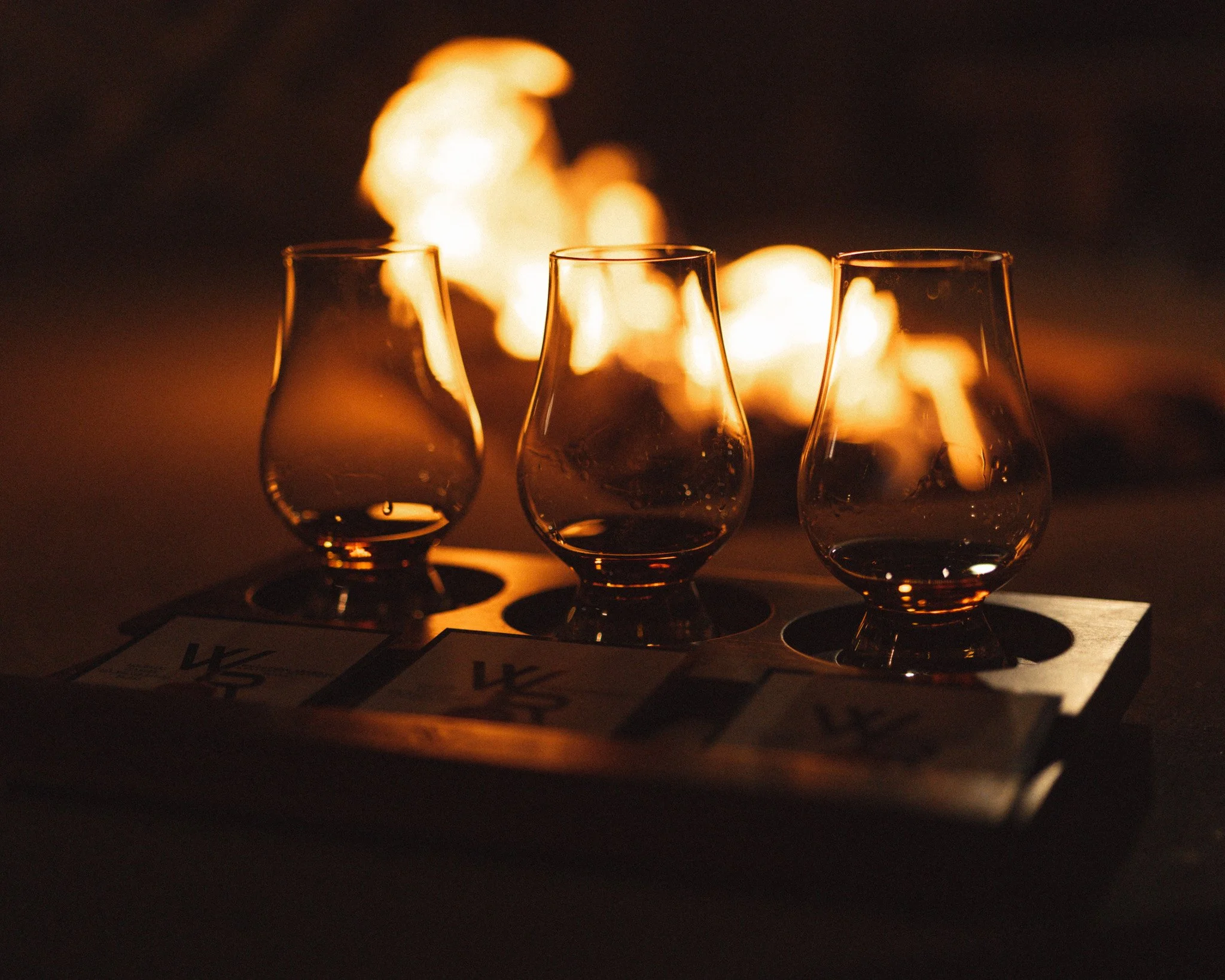Three empty glasses placed on a surface, with a fire or bright light source in the background creating a warm glow and reflections on the glasses.