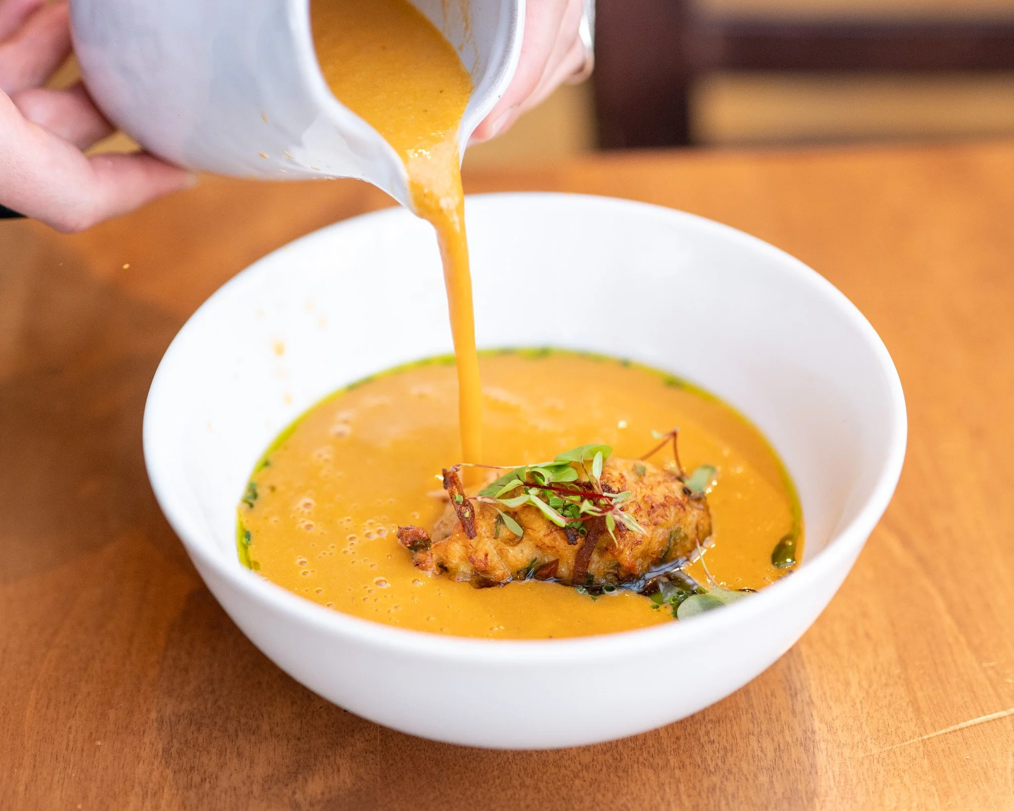 A white bowl of orange soup garnished with microgreens and a fried item, with a hand pouring a creamy orange sauce into it.