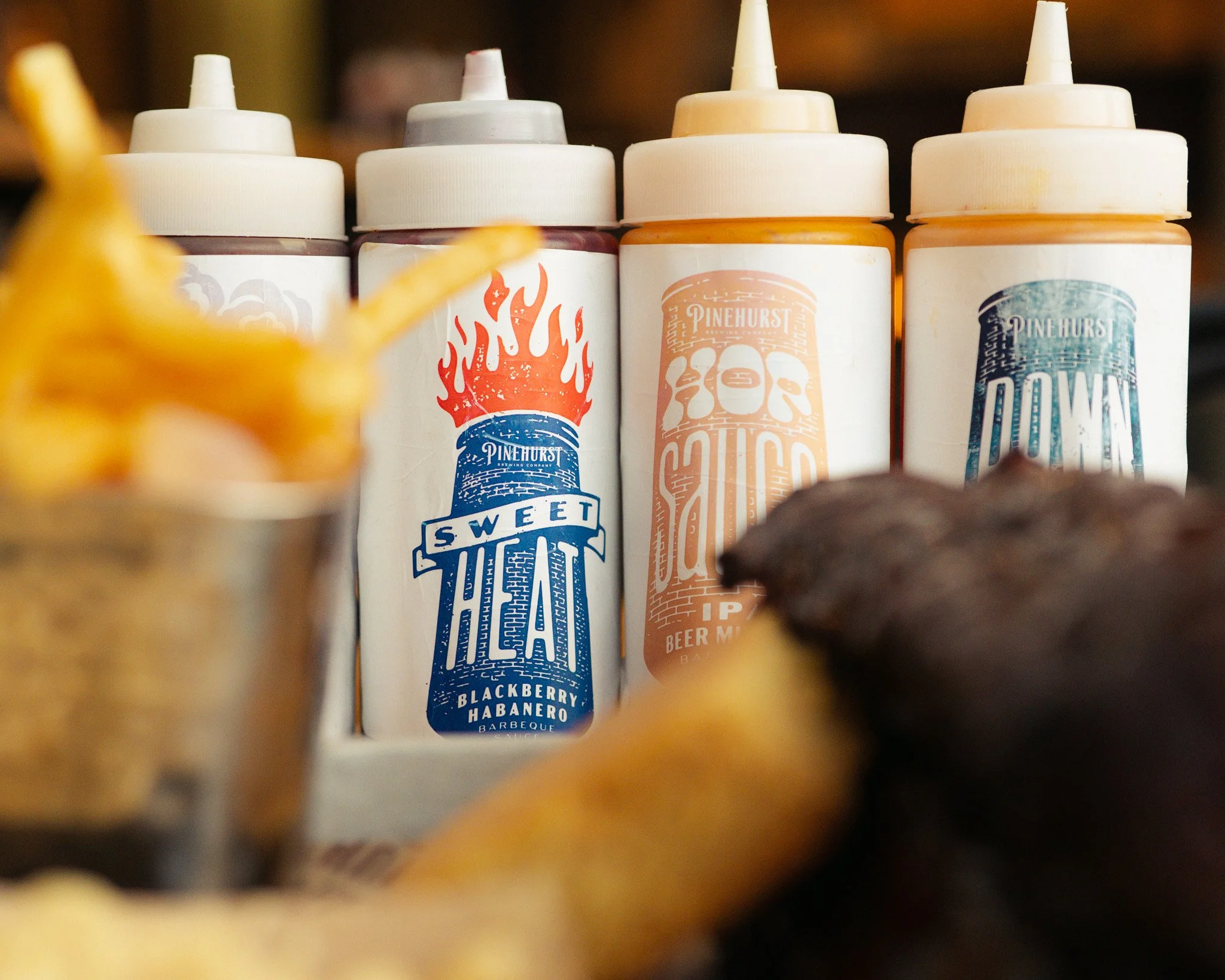Close-up of condiment bottles with labels reading ‘Sweet,’ ‘BBQ,’ ‘Beer Mignon,’ and ‘Smoke,’ with a blurred foreground of what appears to be food or toppings.