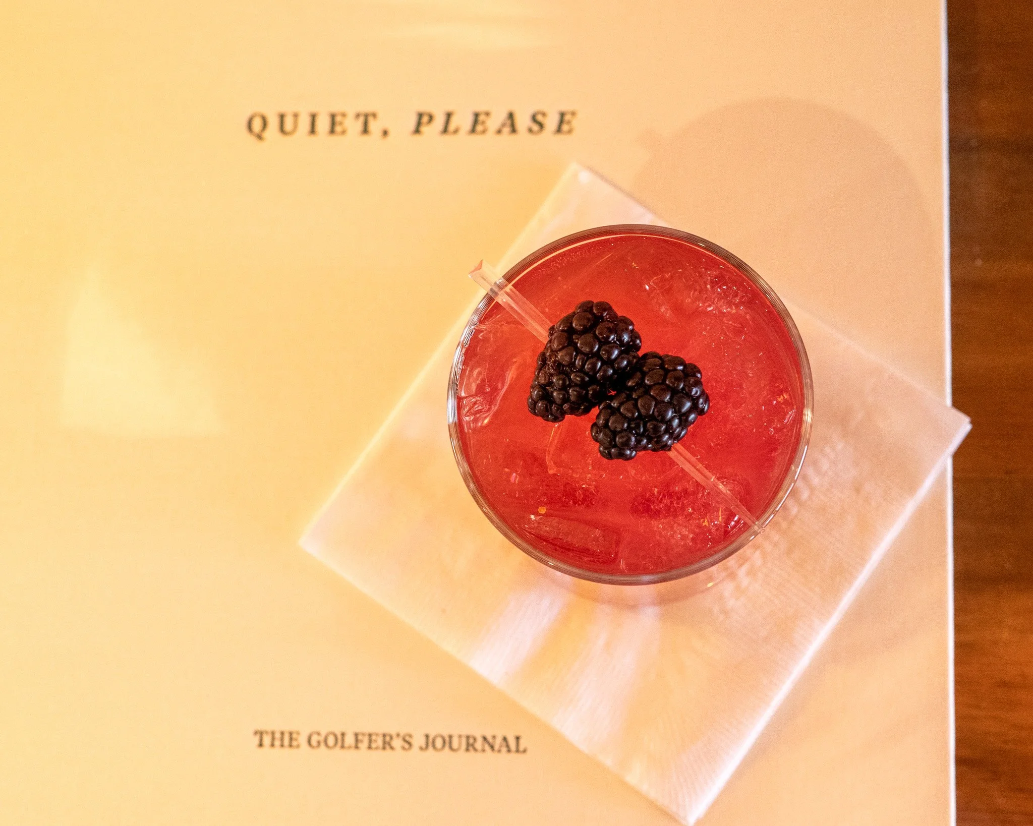 A glass with a red beverage and blackberries on top, placed on a white napkin on a wooden surface with part of a magazine or newspaper visible underneath reading "THE GOLFER'S JOURNAL."