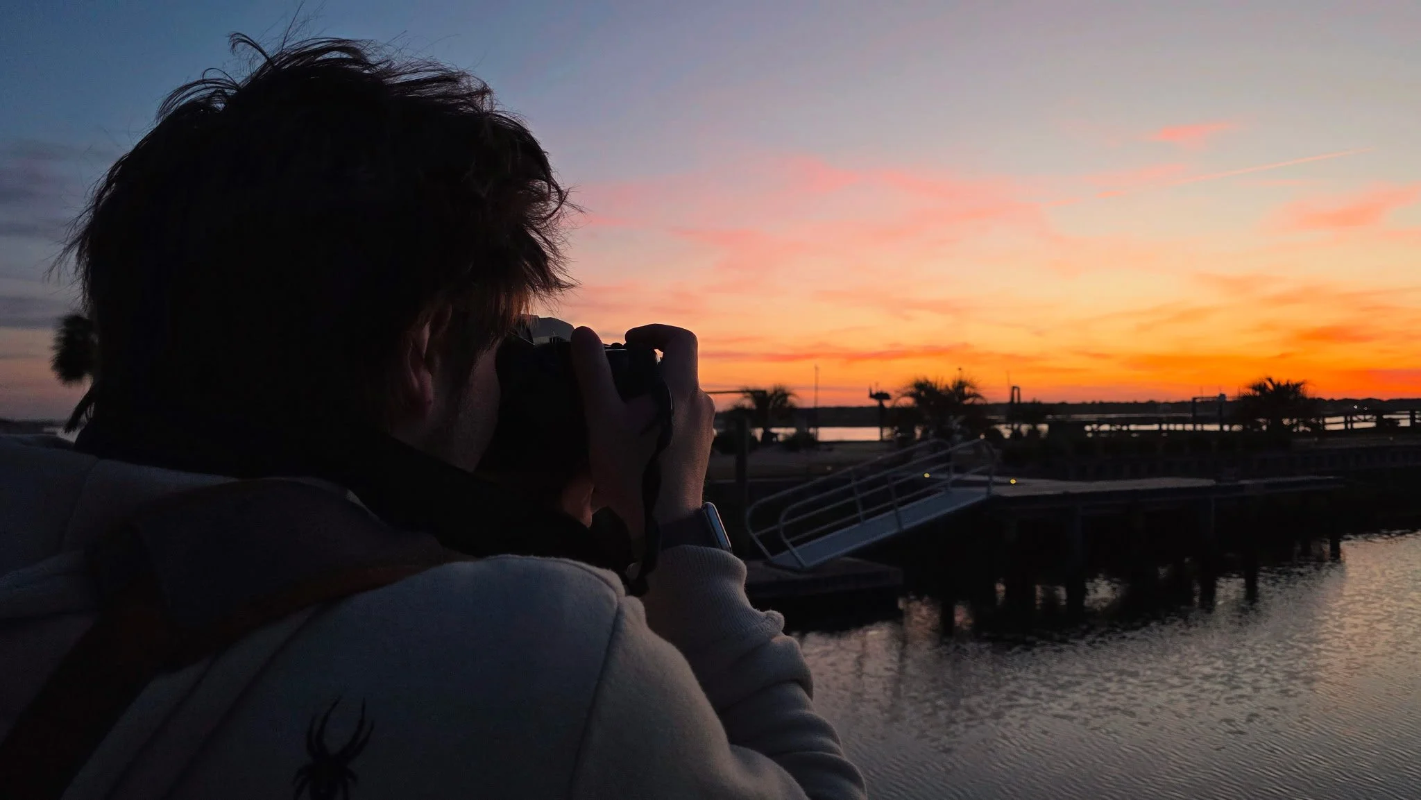 A person with curly hair taking a photograph of a colorful sunset over a waterway with trees and a bridge in the background.